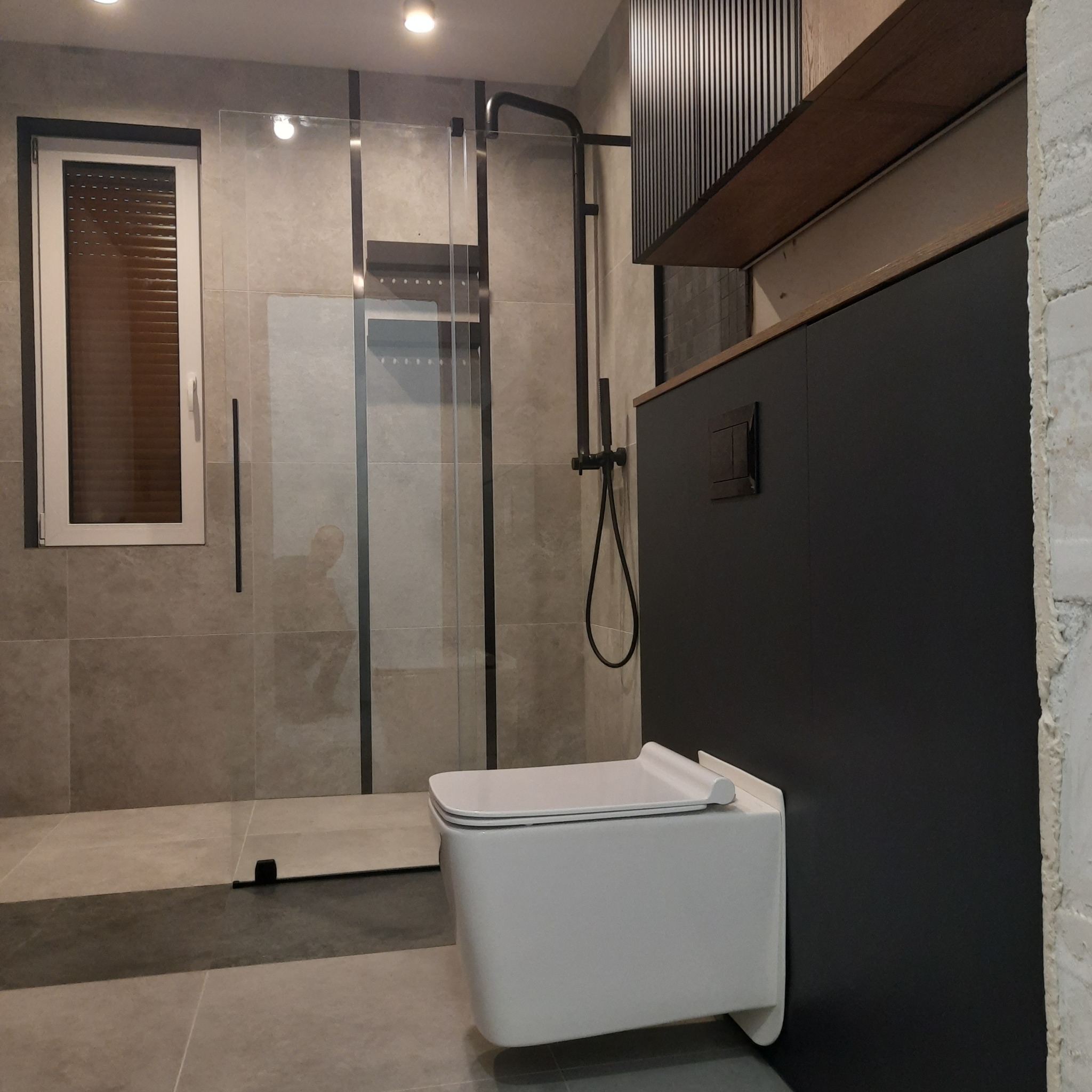 Nowoczesna łazienka z kabiną prysznicową typu walk-in, podwieszaną toaletą i szarą glazurą na ścianach, uzupełniona o czarne akcenty armatury i mebli.