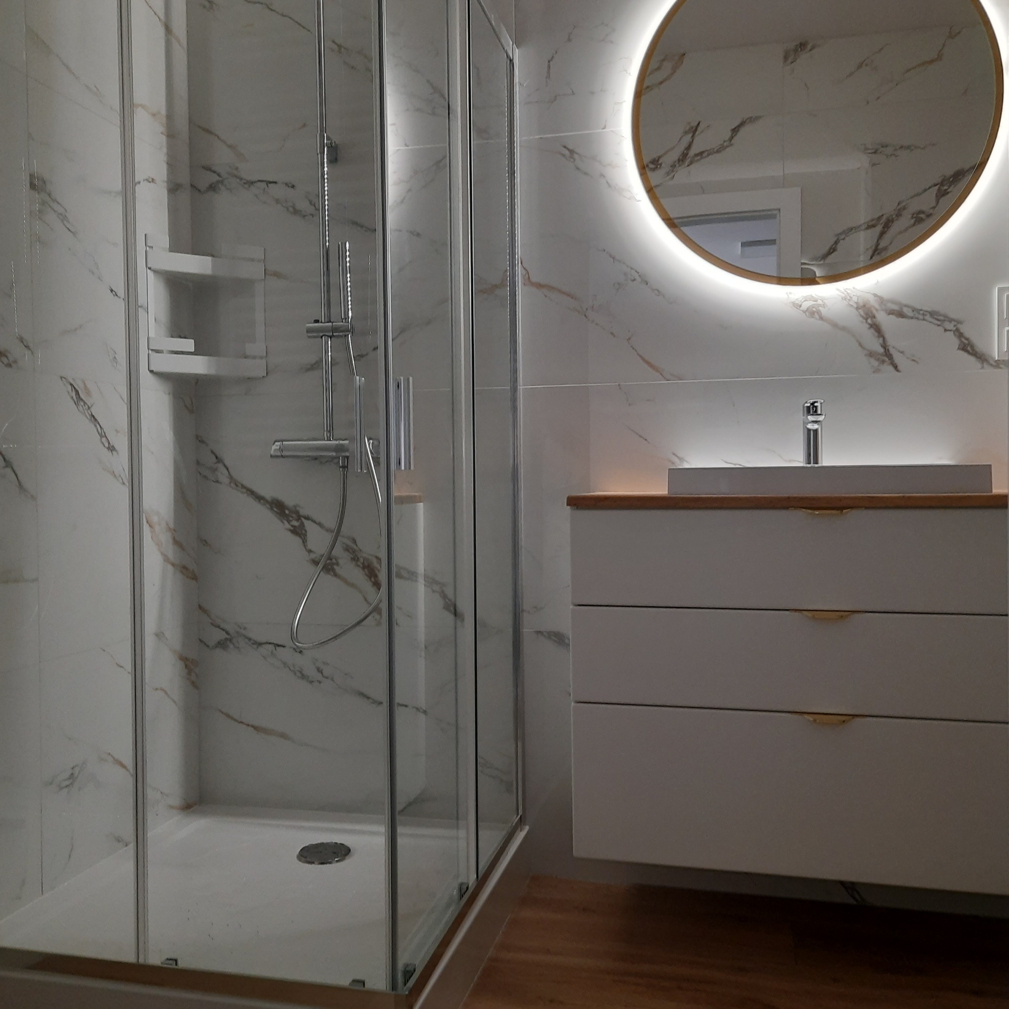 Nowoczesna łazienka z kabiną prysznicową, umywalką na szafce z szufladami i okrągłym lustrem z podświetleniem LED, wykończona płytkami imitującymi marmur z brązowymi żyłami.