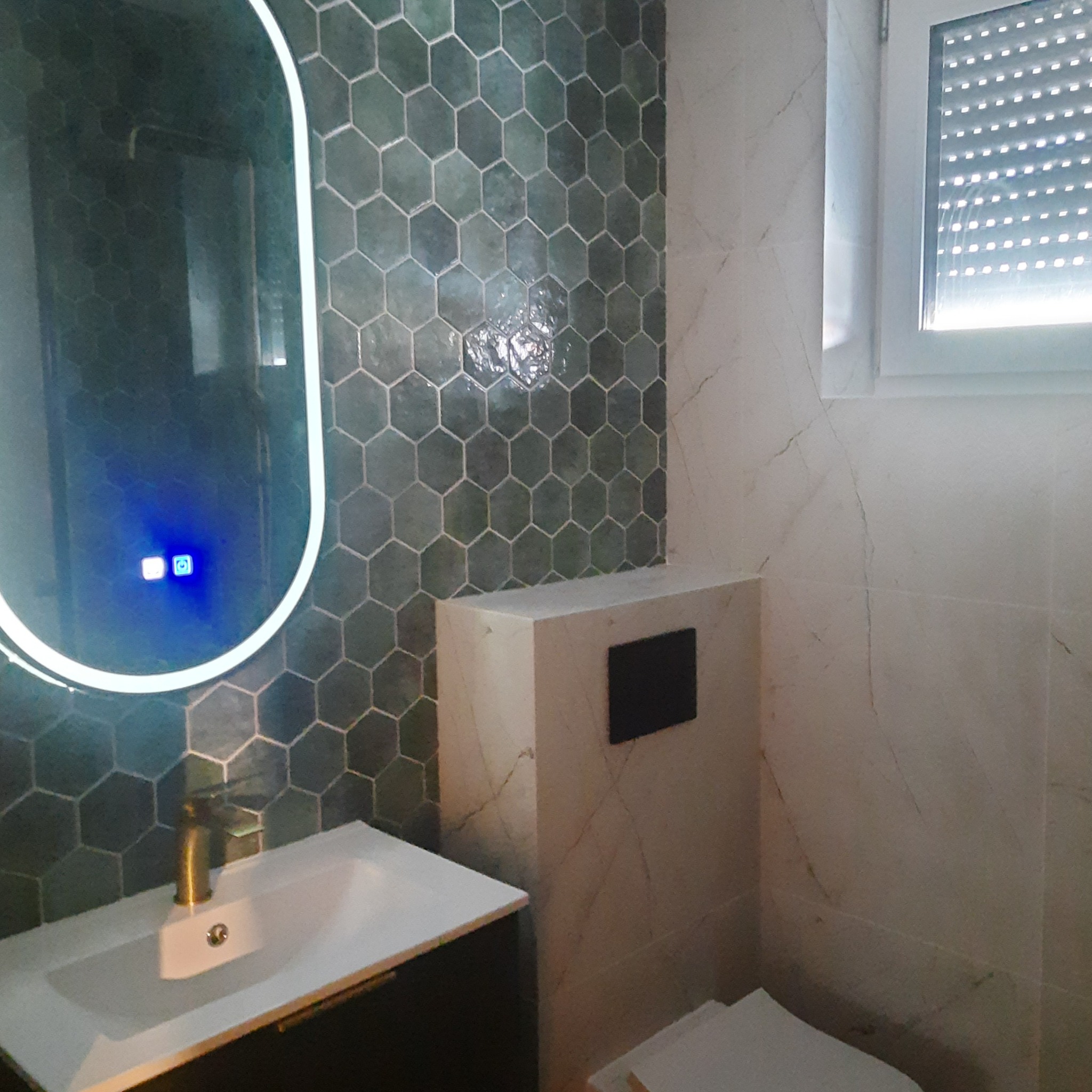 Nowoczesna łazienka z umywalką, podświetlanym lustrem i heksagonalnymi płytkami w odcieniach zieleni oraz marmurowymi płytkami na ścianie, widoczny fragment toalety.