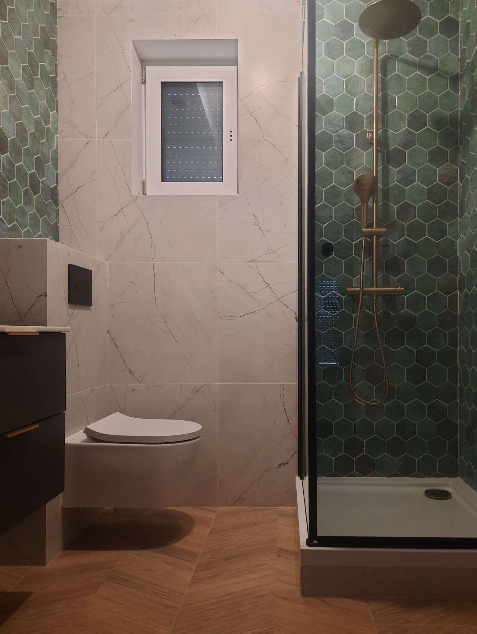 Nowoczesna łazienka z heksagonalnymi płytkami w odcieniach zieleni w strefie prysznicowej, marmurowe płytki na pozostałych ścianach, podwieszana toaleta i podłoga z paneli ułożonych we wzór jodełki.
