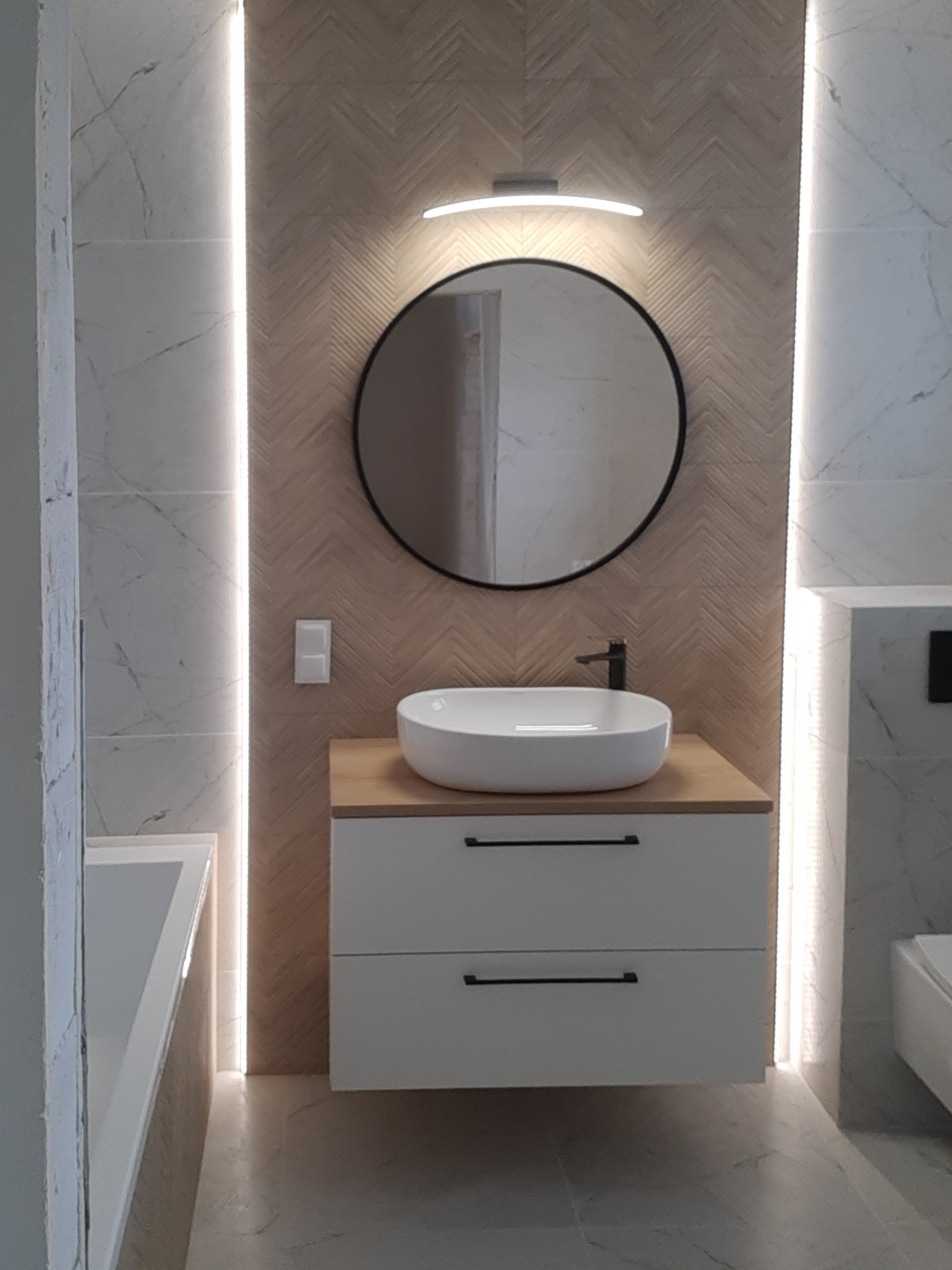 Nowoczesna łazienka z umywalką nablatową, okrągłym lustrem w czarnej ramie i podświetleniem LED wzdłuż krawędzi płytek imitujących marmur.