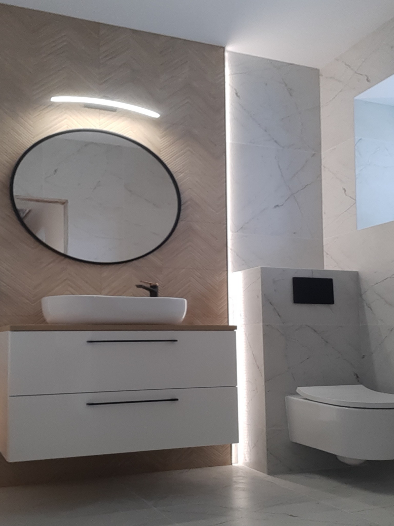 Nowoczesna łazienka z umywalką nablatową, okrągłym lustrem podświetlanym LED, zabudowaną toaletą i płytkami imitującymi drewno jodełkę na ścianie.