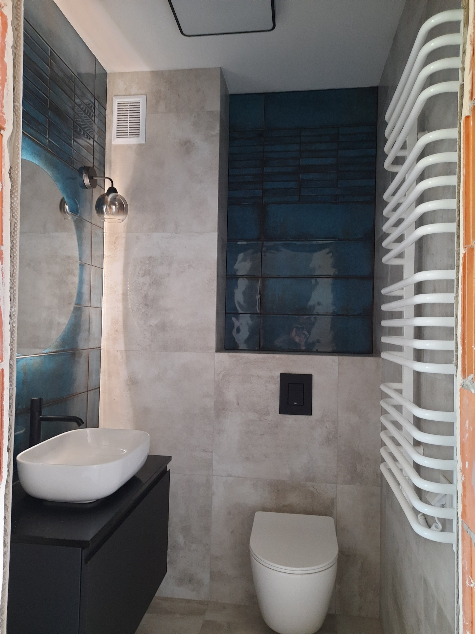 Nowoczesna łazienka z umywalką nablatową na czarnej szafce, podwieszaną toaletą, grzejnikiem drabinkowym i akcentem z ciemnoniebieskich płytek.