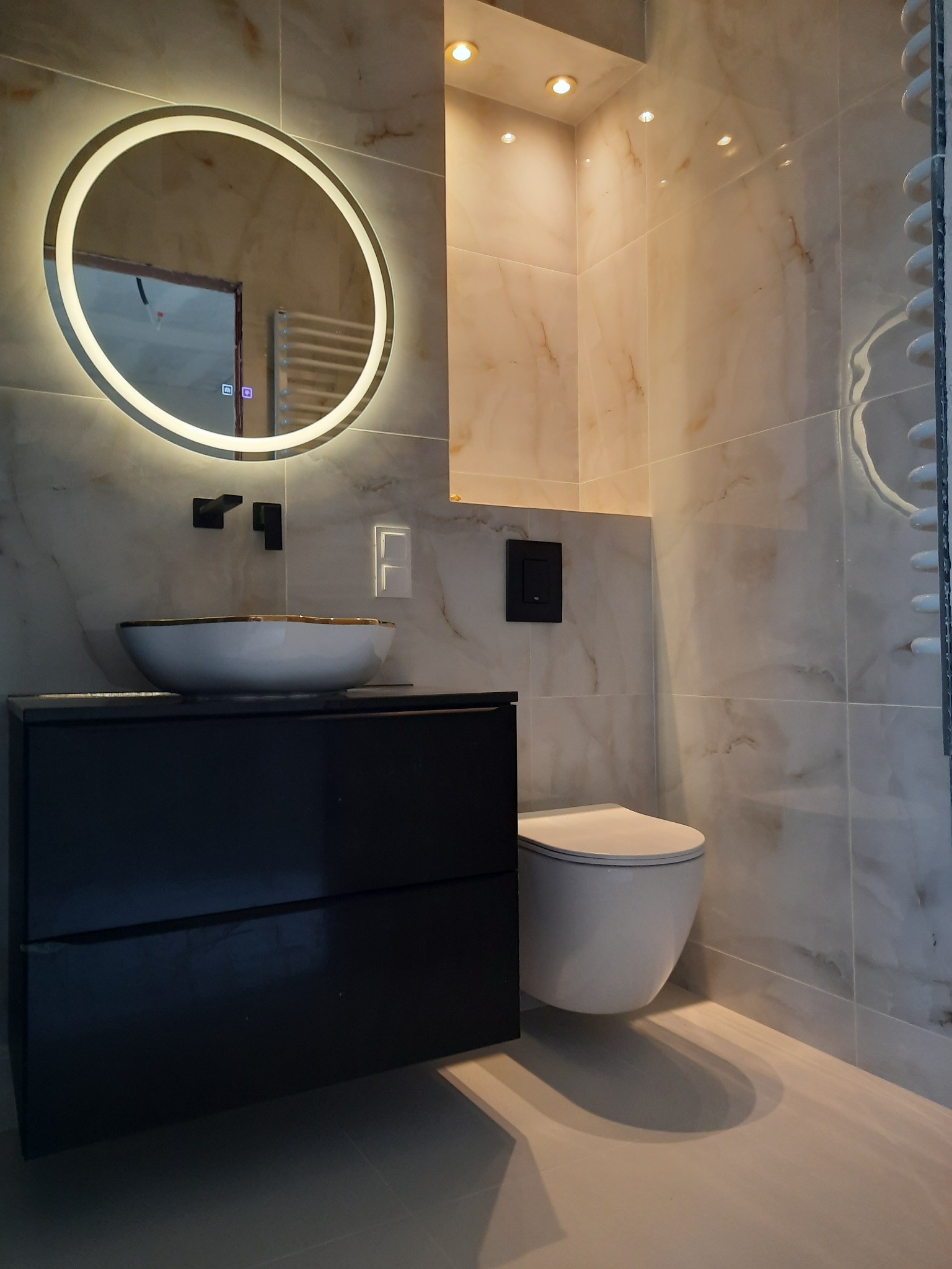 Nowoczesna łazienka z marmurowymi płytkami, okrągłym lustrem z podświetleniem LED, wiszącą toaletą i czarną szafką podumywalkową.