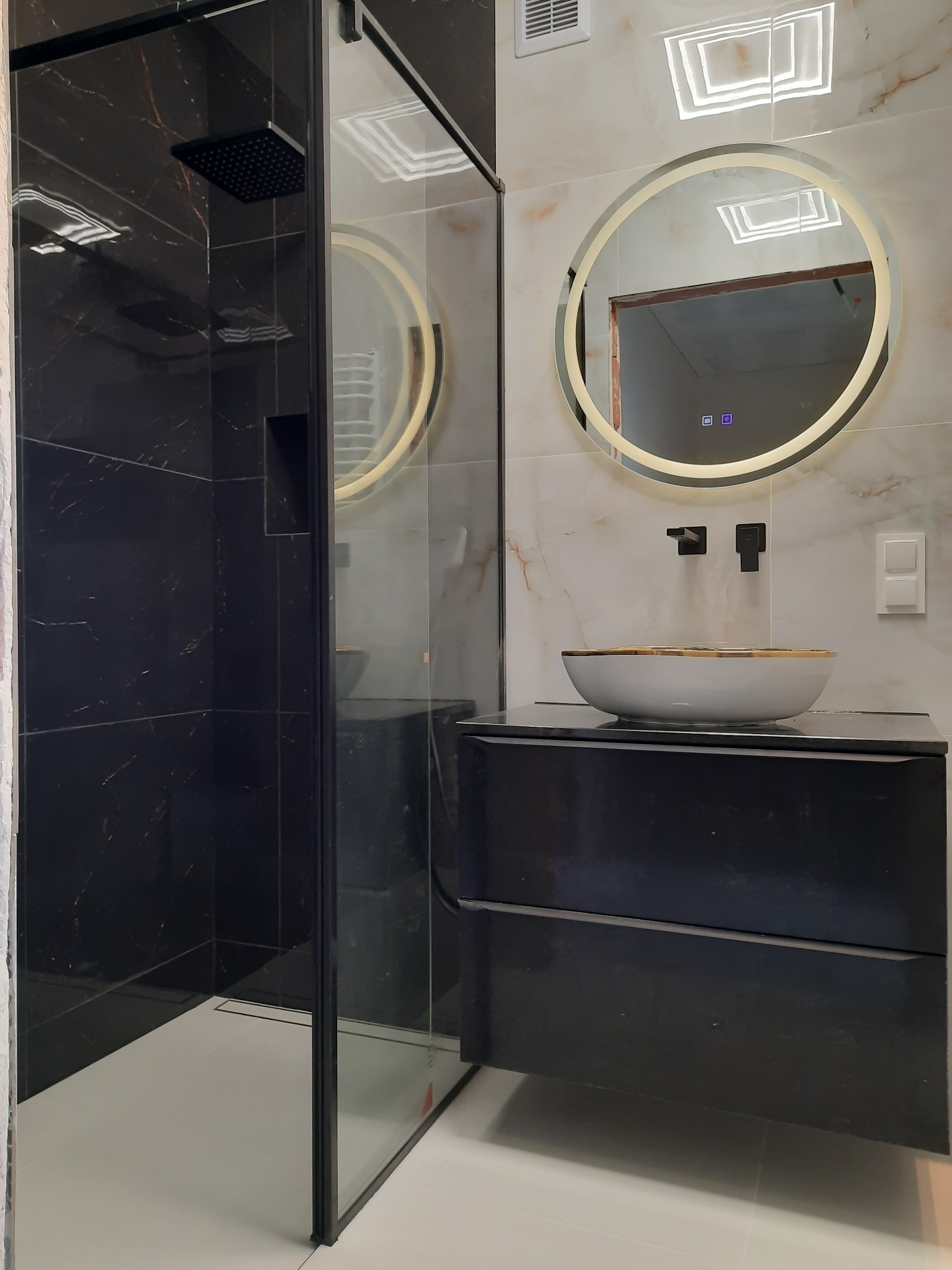 Nowoczesna łazienka z czarnymi płytkami imitującymi marmur w kabinie prysznicowej typu walk-in, okrągłym lustrem z podświetleniem LED nad białą umywalką z czarną szafką i czarną armaturą.
