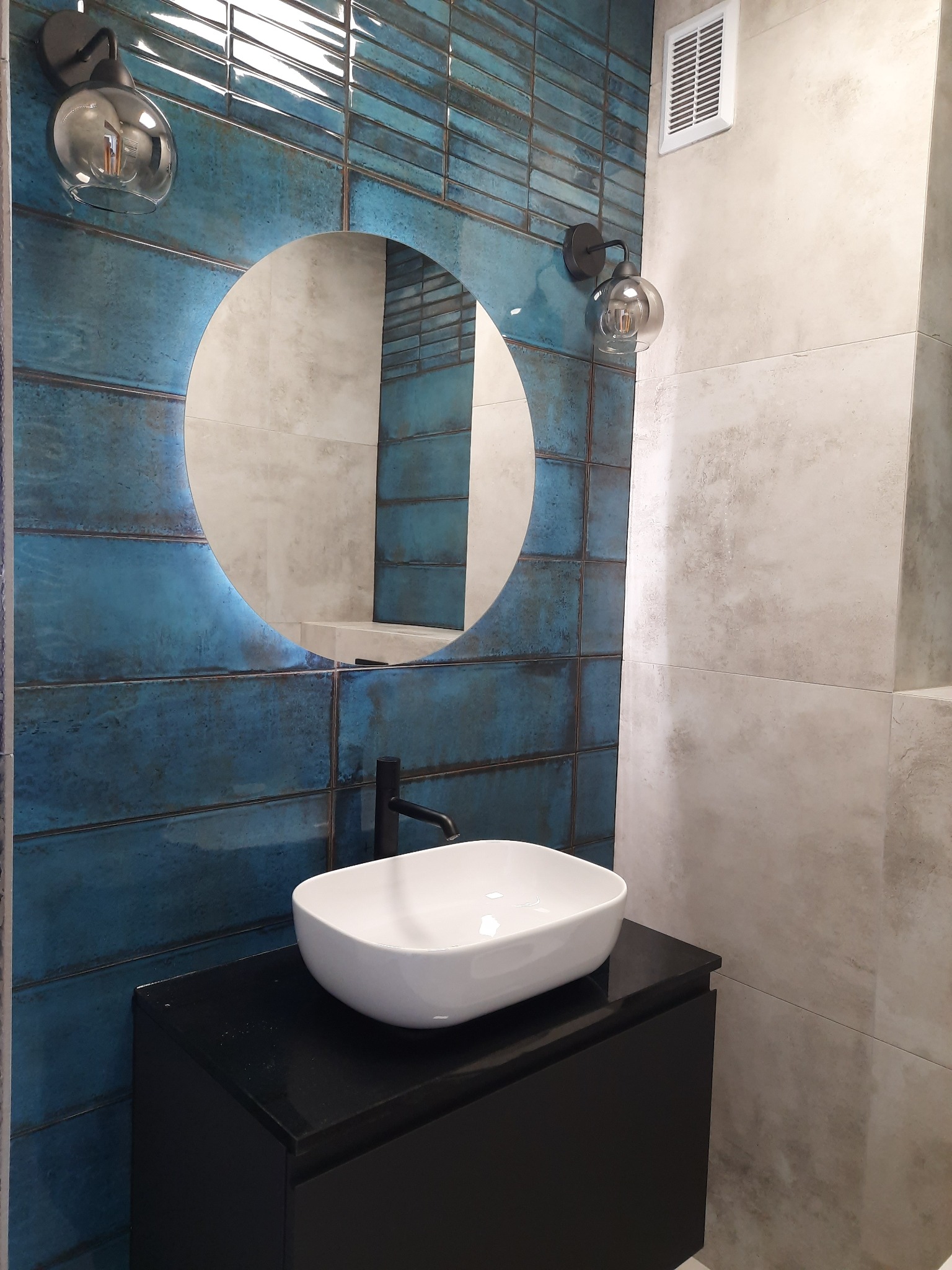 Nowoczesna łazienka z umywalką nablatową, okrągłym lustrem z podświetleniem LED, turkusowymi płytkami imitującymi cegłę i szklanymi lampami ściennymi.