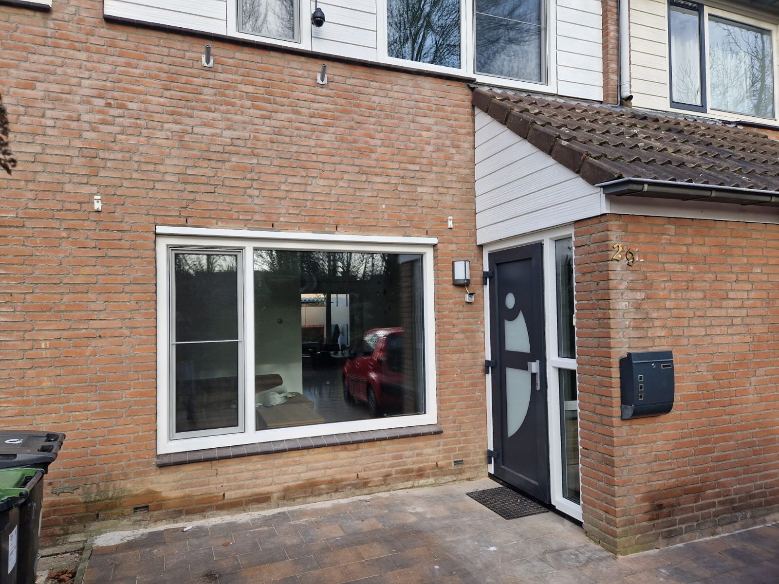 Ceglana fasada domu z nowym oknem z białymi ramami, ciemnymi drzwiami wejściowymi z ozdobnym szkleniem i skrzynką na listy. W oknie odbija się wnętrze z czerwonym samochodem.