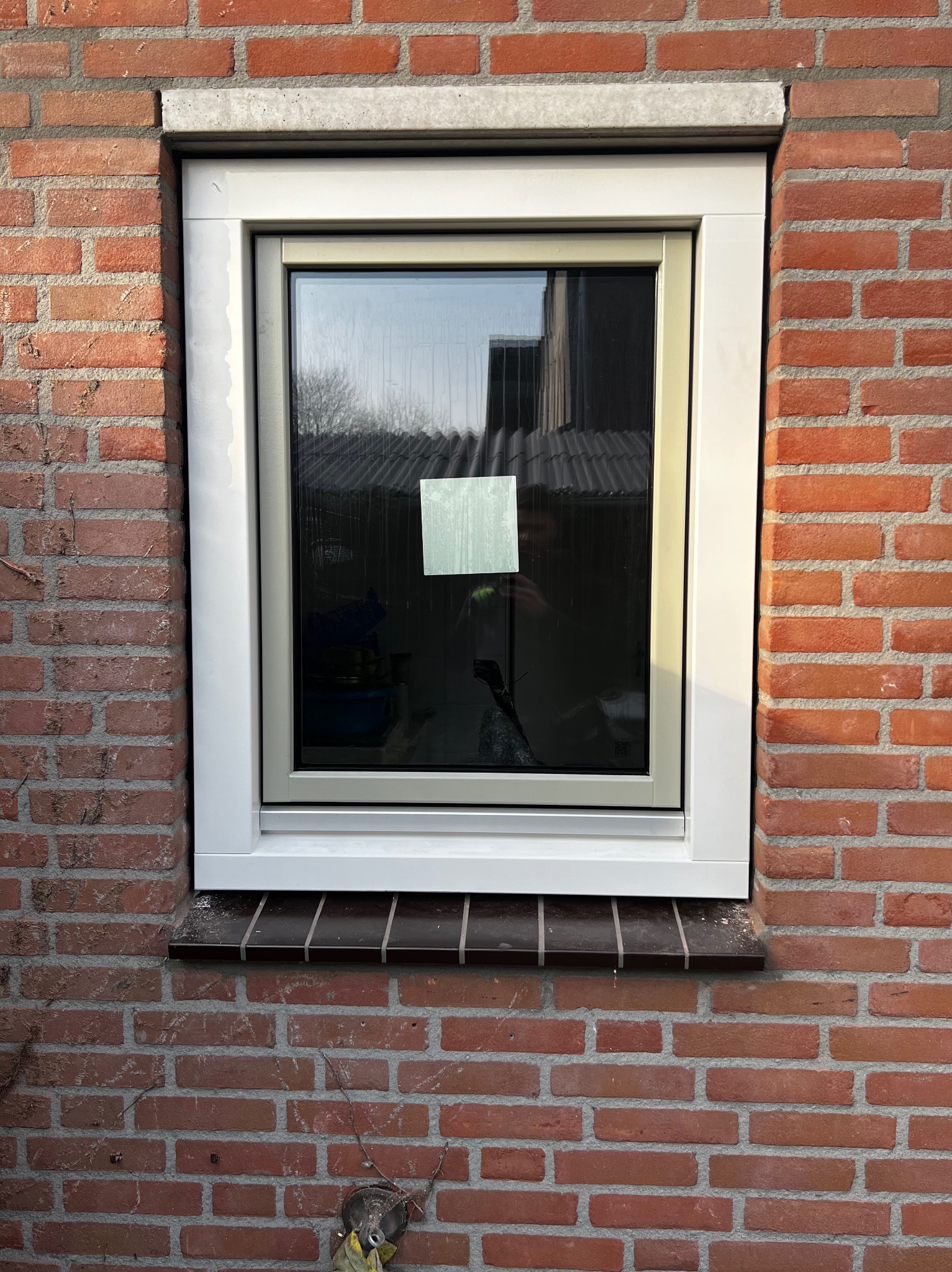 Nowe, białe okno PCV zamontowane w ścianie z czerwonej cegły, widoczny parapet zewnętrzny z ciemnego materiału.