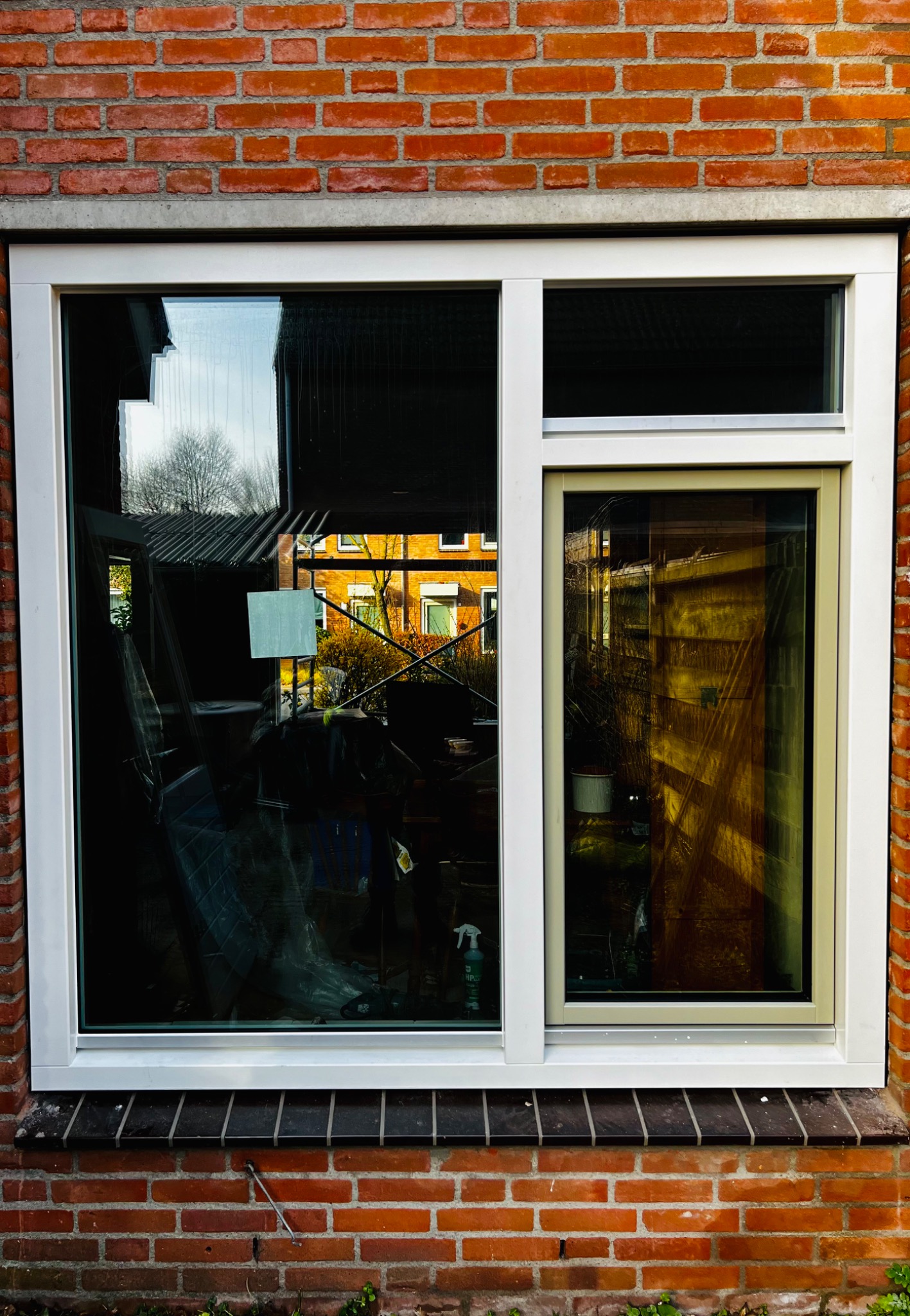 Nowe okno PCV z białymi ramami w murze z czerwonej cegły, widoczne odbicie otoczenia w szybach i częściowo zasłonięte wnętrze pomieszczenia.