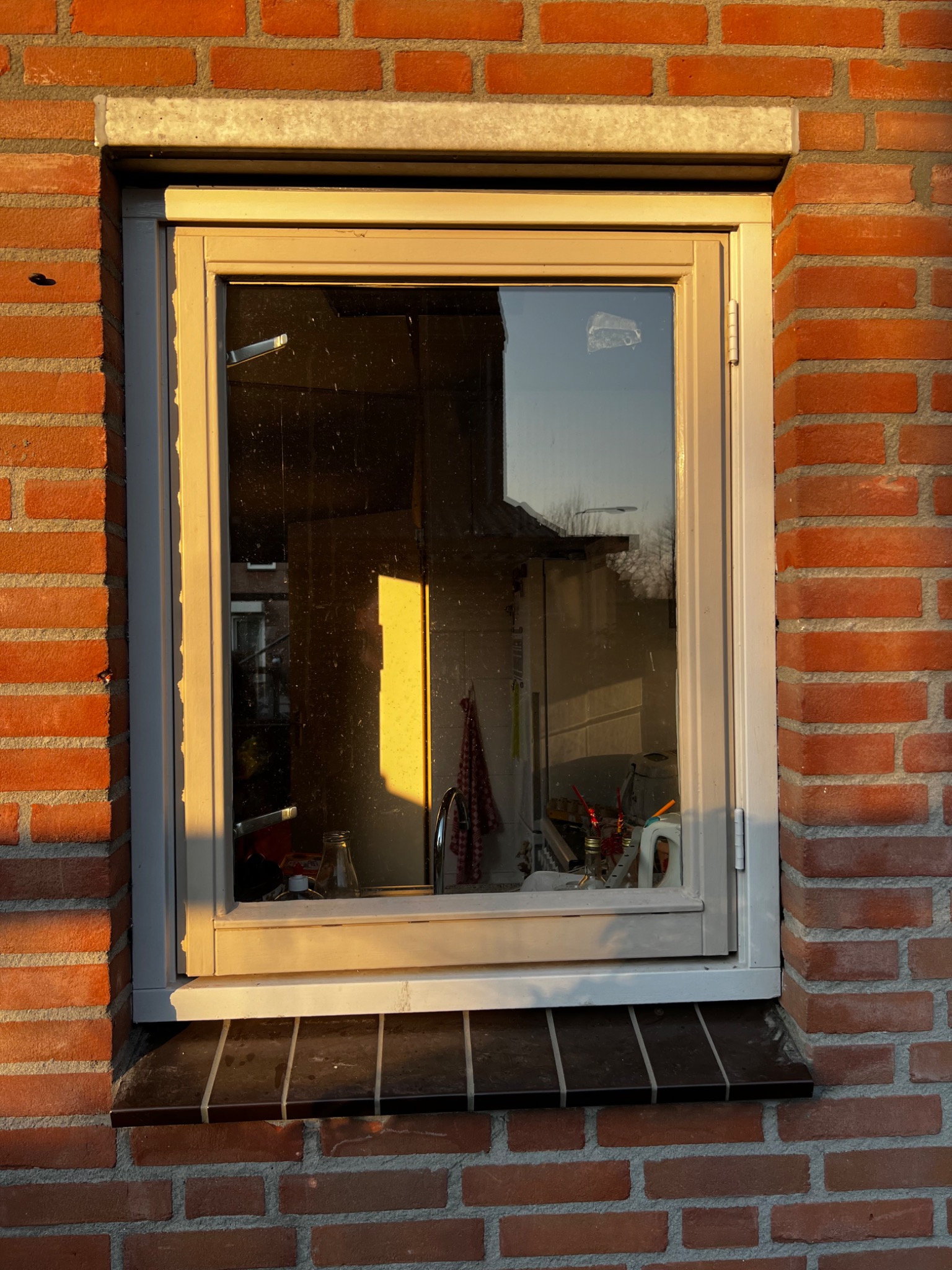 Zamknięte okno z beżową ramą w czerwonej ceglanej ścianie, z widocznymi refleksami zachodzącego słońca na szybie i ścianie, oraz widocznym fragmentem wnętrza kuchni w odbiciu.