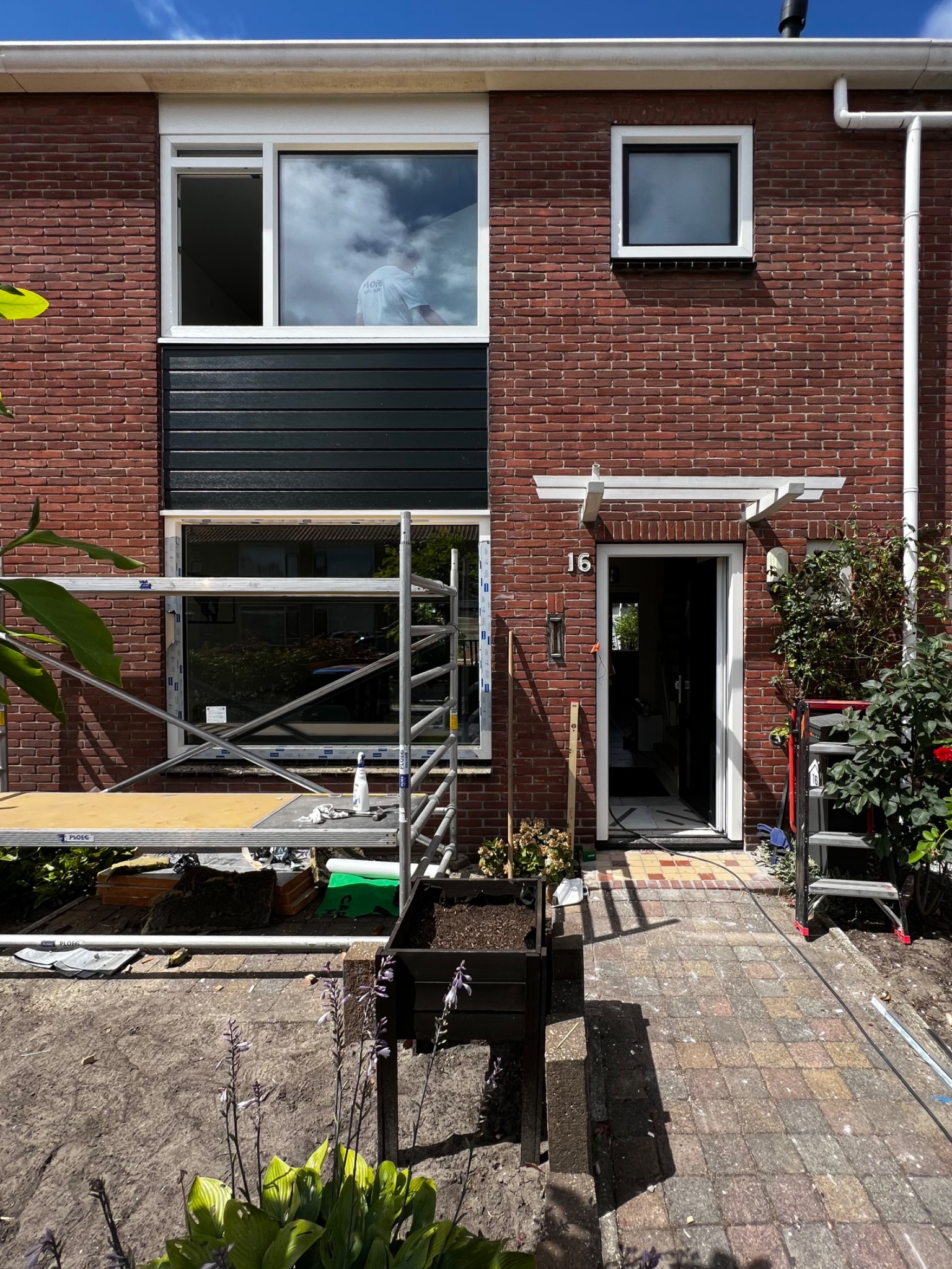 Dom jednorodzinny z czerwonej cegły w trakcie remontu; widoczne rusztowanie przed oknem, otwarte drzwi wejściowe, numer domu 16.