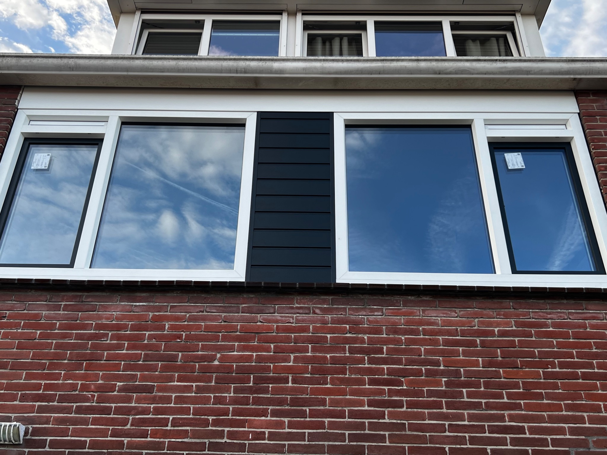 Ceglana fasada domu z nowymi oknami w białych ramach, pomiędzy oknami pionowy panel z czarnych desek.