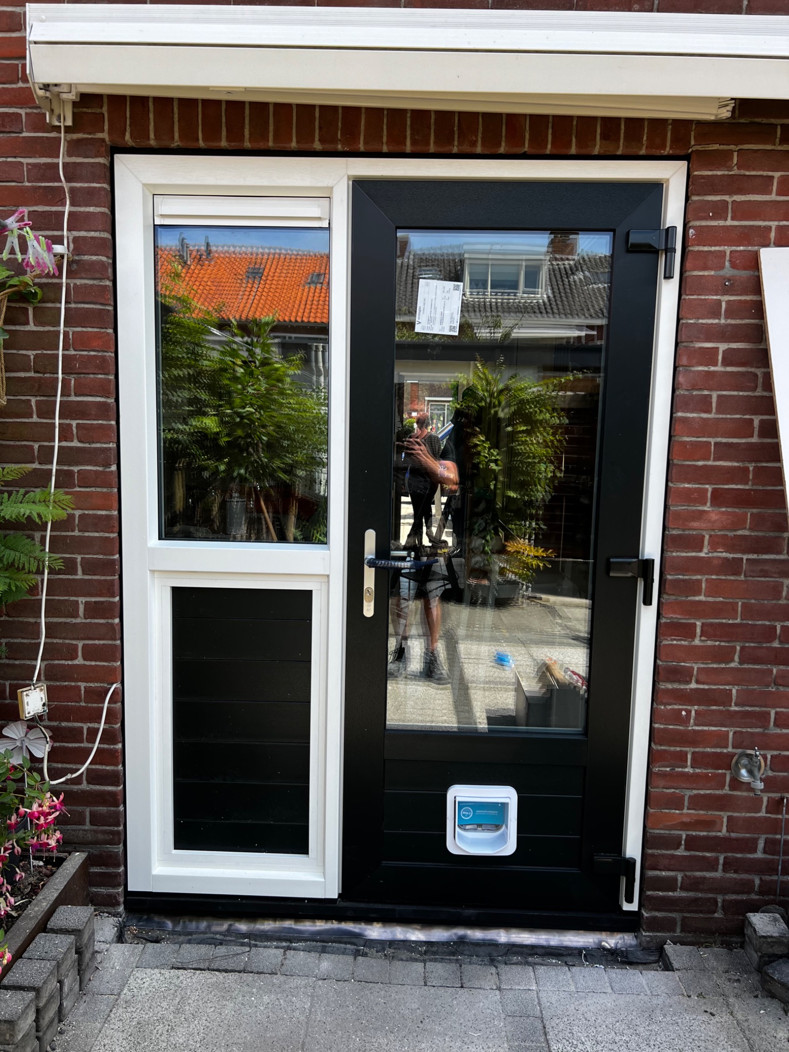 Nowoczesne drzwi zewnętrzne z czarną ramą, białym obramowaniem i oknem, zamontowane w ceglanej ścianie, z widocznym przejściem dla zwierząt i odbiciem fotografa w szybie.