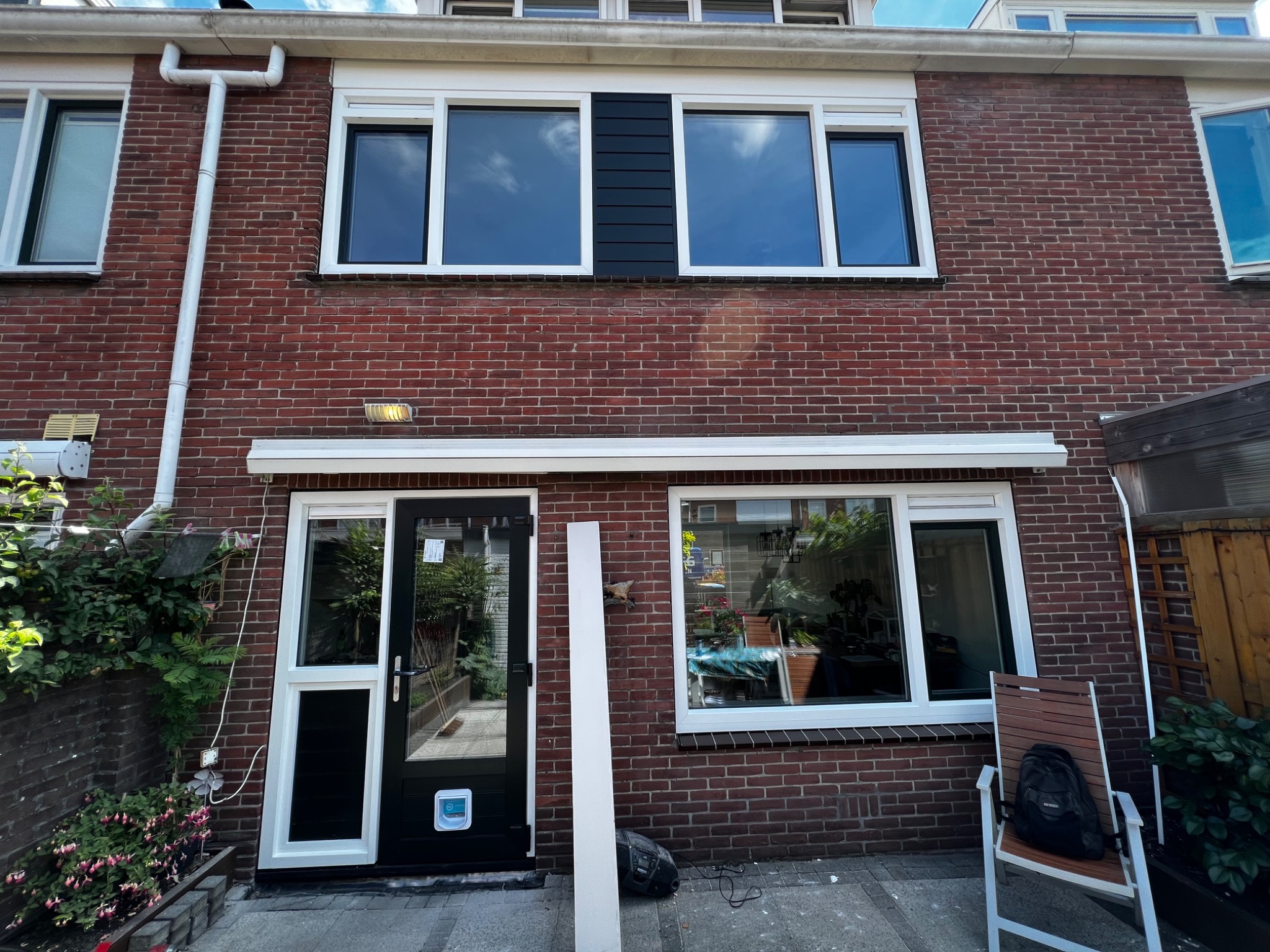 Ceglana elewacja dwukondygnacyjnego domu z białymi ramami okiennymi i drzwiami, widoczny fragment rynny i biały panel elewacyjny oparty o ścianę przy drzwiach.