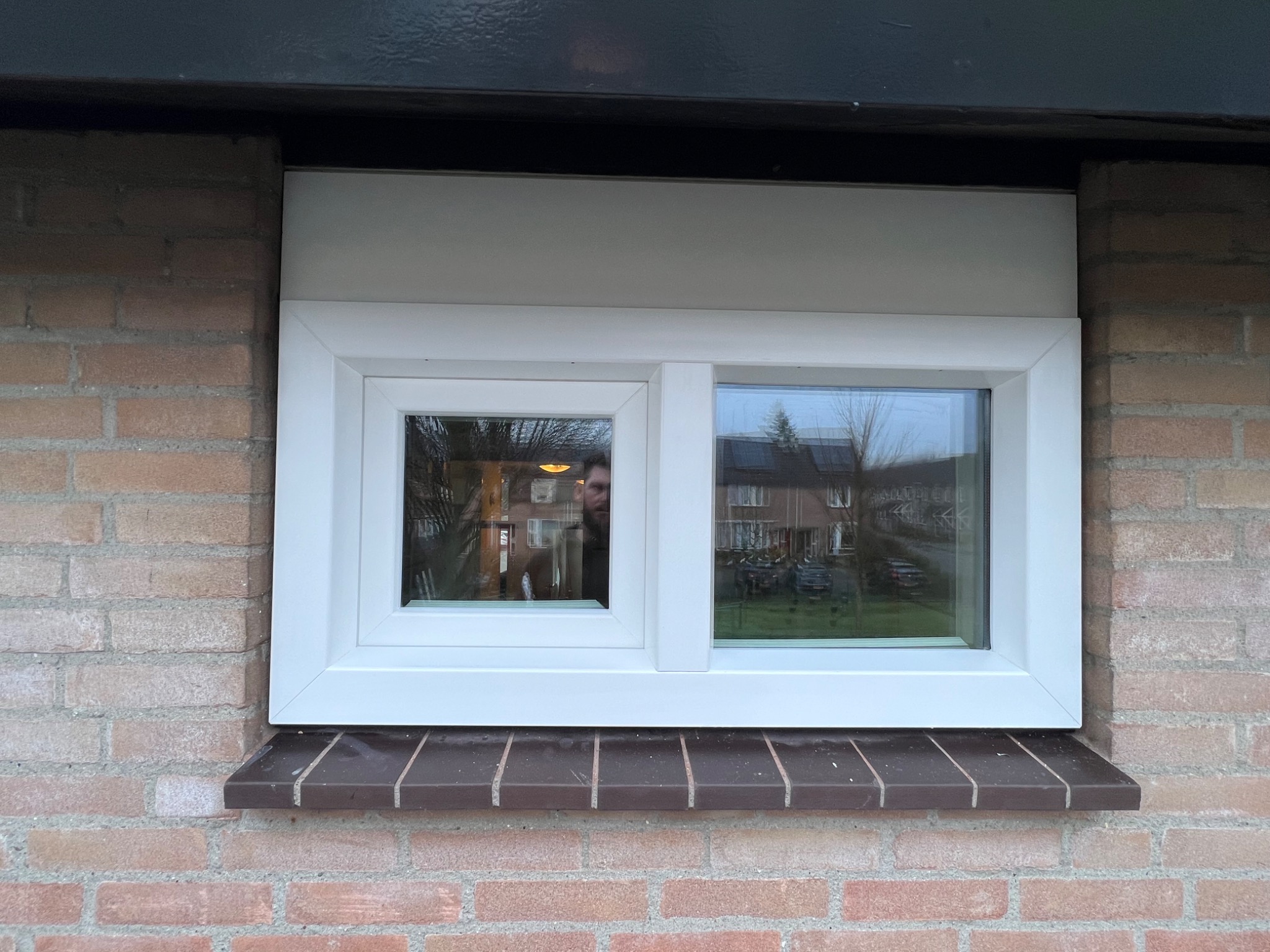 Białe, dwudzielne okno PCV wmurowane w ścianę z cegły klinkierowej, z widocznym odbiciem osoby w szybie.