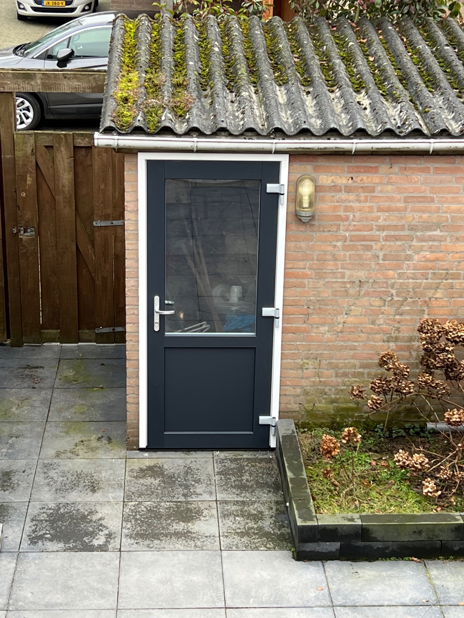 Nowoczesne, grafitowe drzwi zewnętrzne z białym obramowaniem, zamontowane w murze z czerwonej cegły, z widoczną lampą zewnętrzną i porośniętym mchem dachem.
