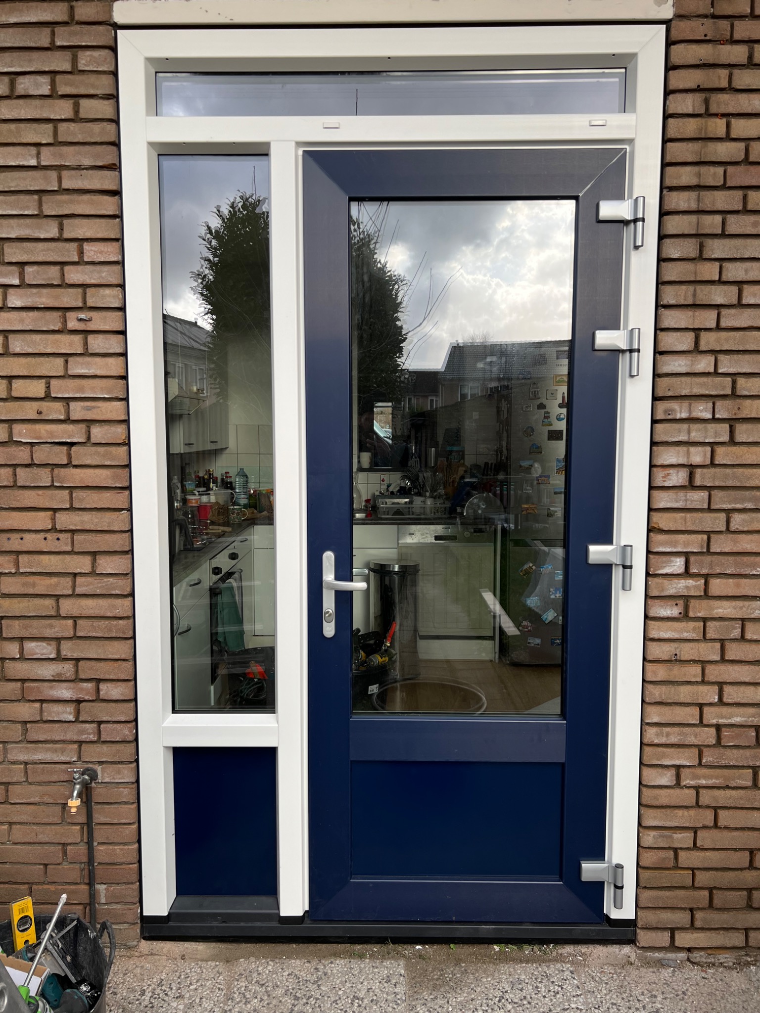 Zewnętrzne drzwi wejściowe z bocznym panelem i górnym oknem, w kolorze białym i granatowym, zamontowane w ścianie z cegły. Przez szybę widoczny fragment kuchni z białymi szafkami i wyposażeniem.