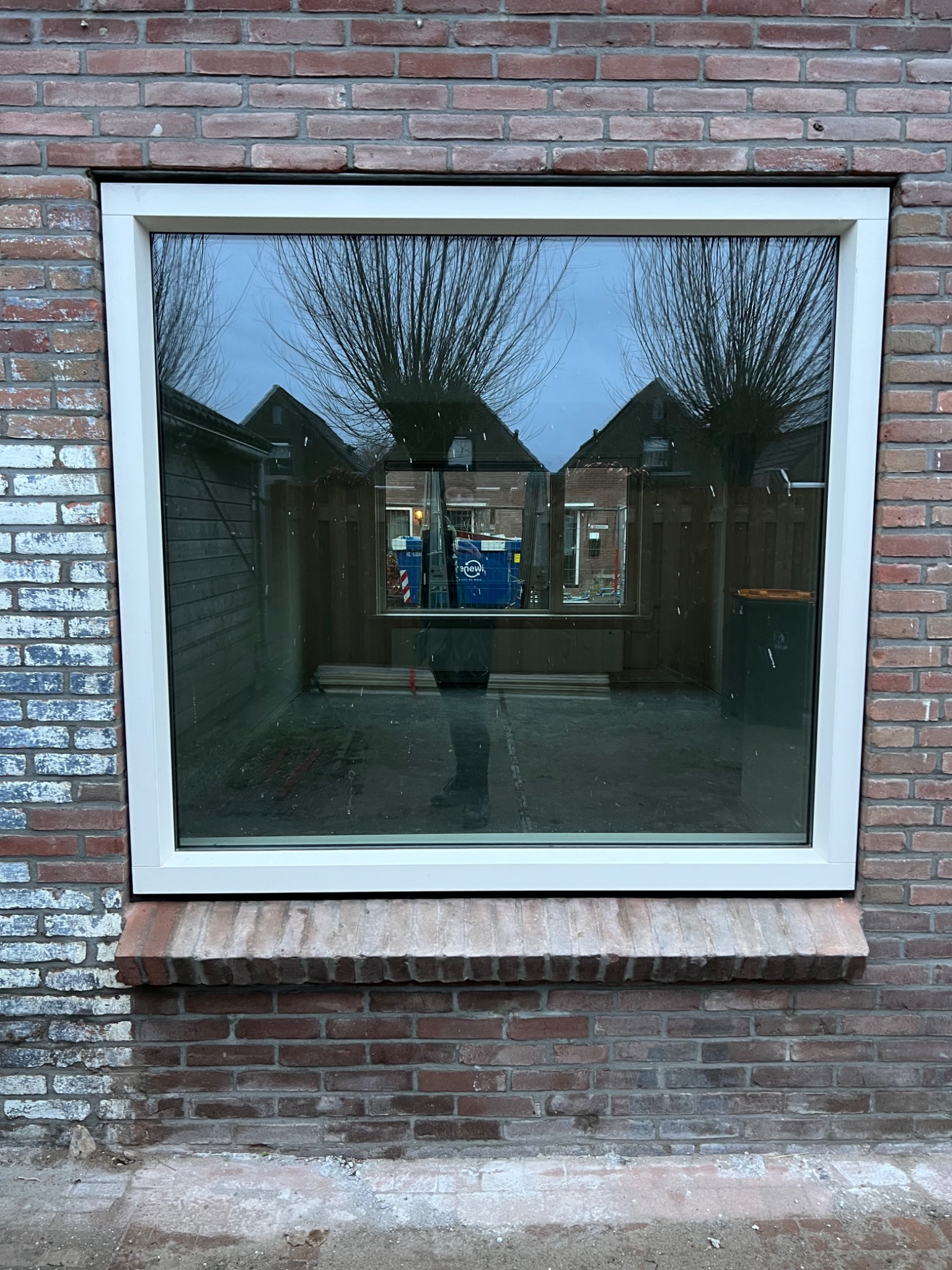 Nowe okno PCV z białą ramą osadzone w ścianie z czerwonej cegły; widoczne odbicie drzew i budynków w szybie.