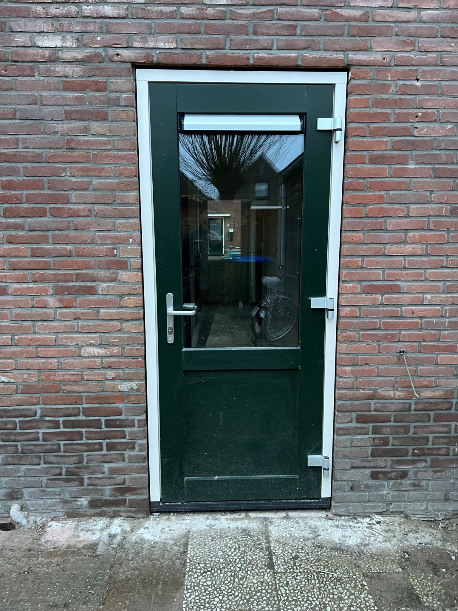 Zewnętrzne drzwi wejściowe, pomalowane na zielono z białymi ramami, osadzone w ścianie z czerwonej cegły, widok na rower wewnątrz budynku przez szybę.