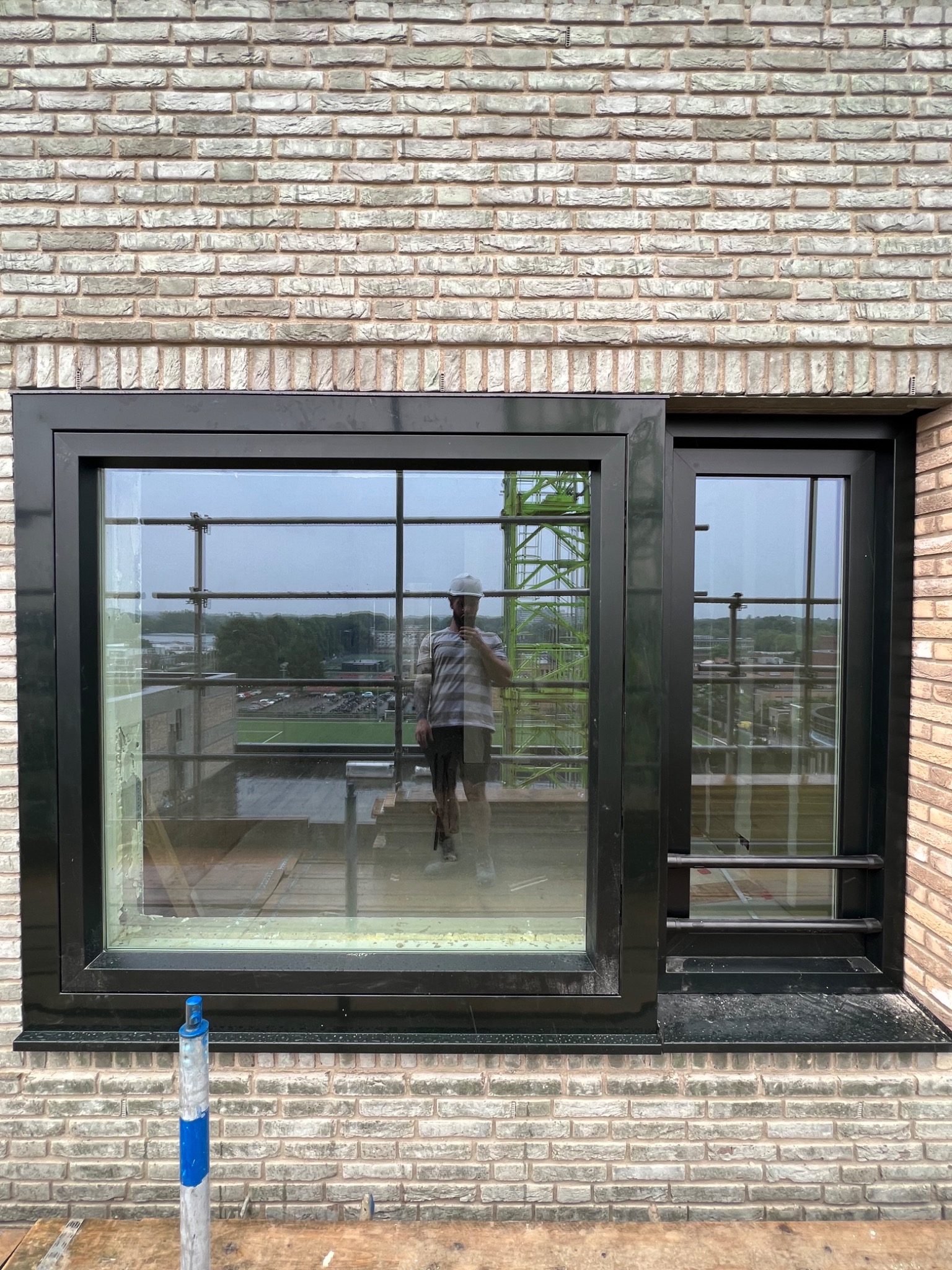 Nowe okno z czarną ramą, zamontowane w ścianie z jasnej cegły. W odbiciu szyby widać pracownika w kasku i fragment żurawia budowlanego.