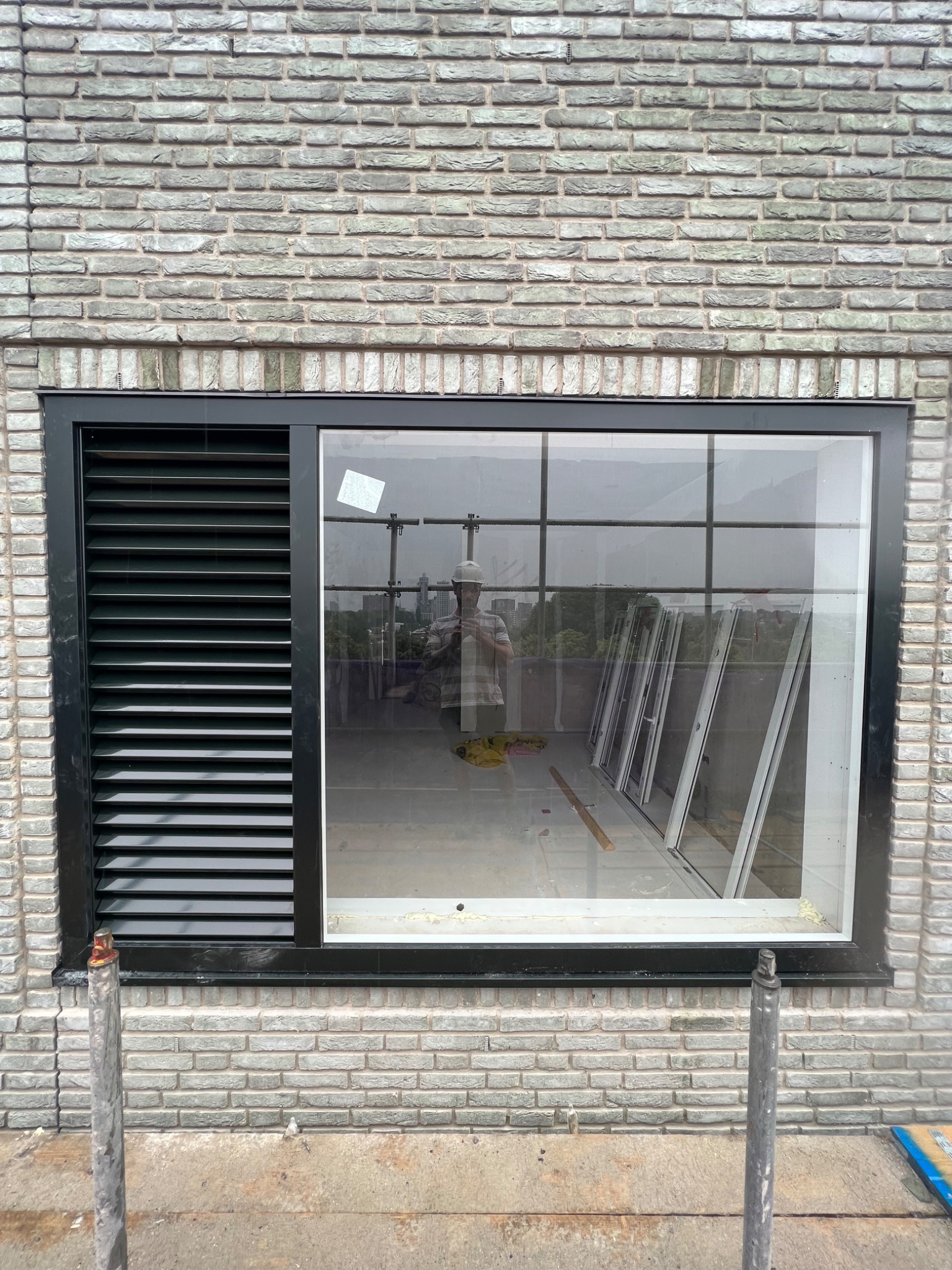 Nowe okno z czarną ramą i żaluzjami, w trakcie montażu w ścianie z szarej cegły, widoczne elementy rusztowania i odbicie osoby w oknie.