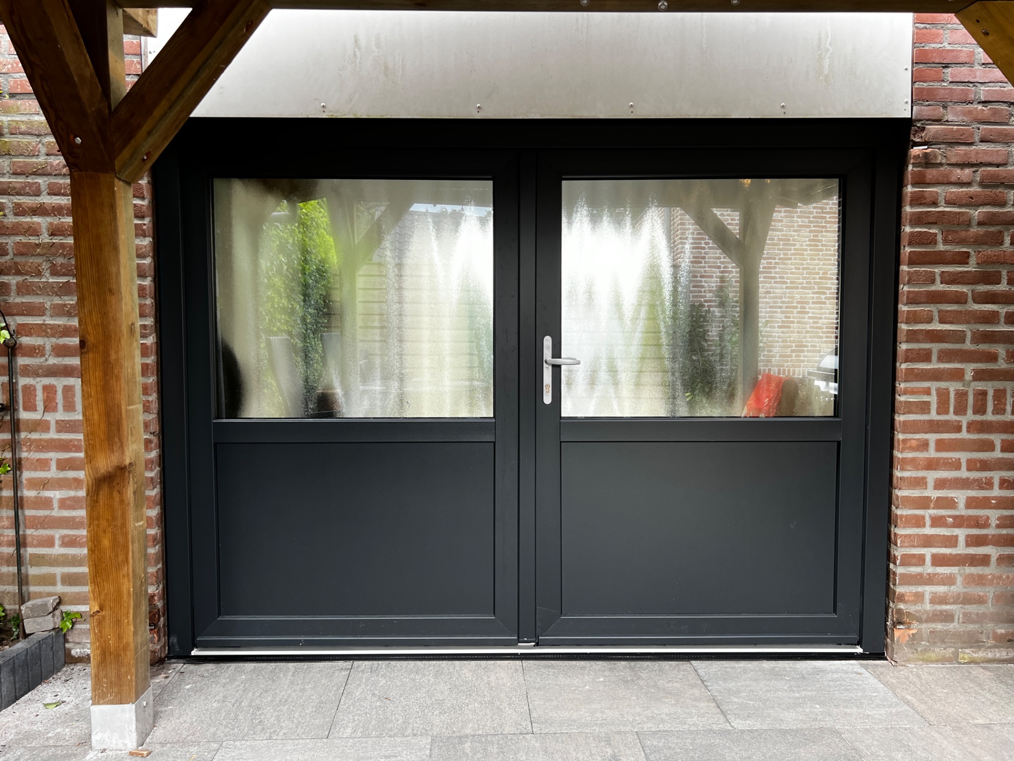 Zamknięte, grafitowe dwuskrzydłowe drzwi z szybami z widokiem na ogród, osadzone w ceglanej ścianie i drewnianej konstrukcji zadaszenia.