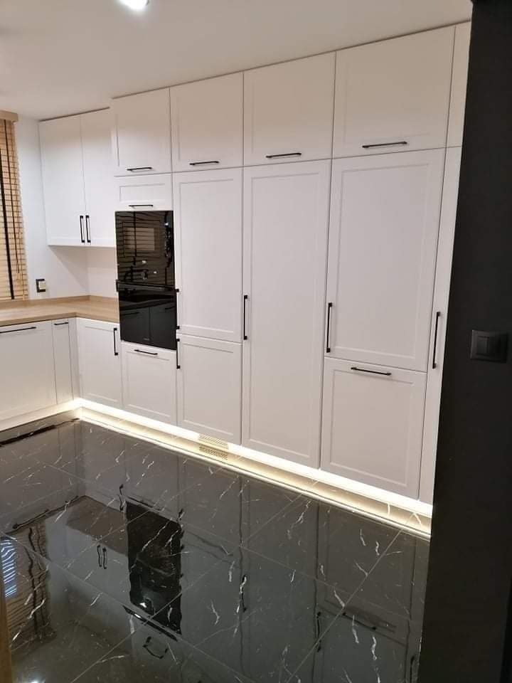 Nowoczesna kuchnia z białymi frontami, czarnymi, podłużnymi uchwytami, piekarnikiem w zabudowie i marmurową podłogą z podświetleniem LED u dołu szafek.