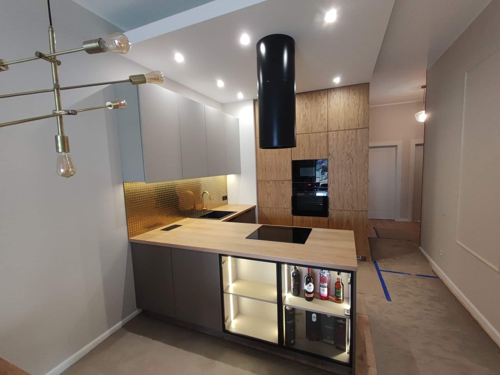 Nowoczesna kuchnia z szarymi szafkami, złotą mozaiką na ścianie, drewnianymi frontami i wyspą z podświetlaną witryną na alkohol, czarny okrągły okap i designerska lampa wisząca.