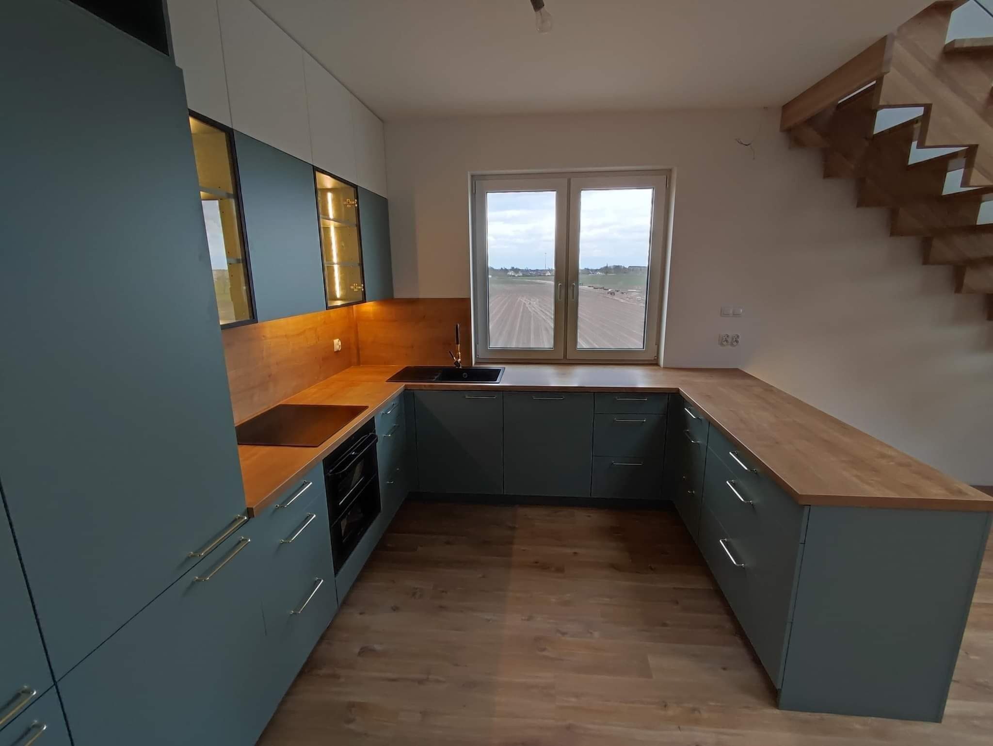 Nowoczesna kuchnia w odcieniach szarości z drewnianym blatem, oświetleniem podszafkowym i widokiem na pole za oknem.