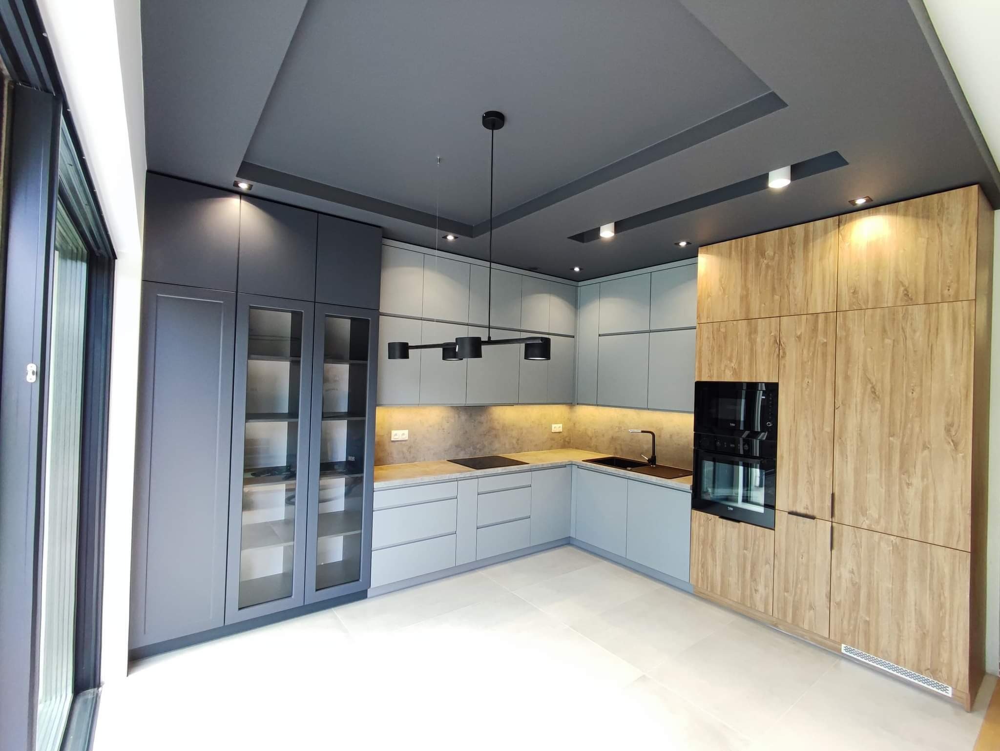 Nowoczesna kuchnia z szarymi szafkami i drewnopodobnym wykończeniem, oświetlenie podszafkowe, czarna armatura i lampa sufitowa.