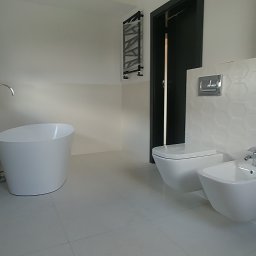 Kamil Kwiatkowski - Nowoczesna, minimalistyczna łazienka z wolnostojącą wanną, wiszącą toaletą i bidetem, białe płytki z heksagonalnym wzorem na ścianie, czarne drzwi w tle.