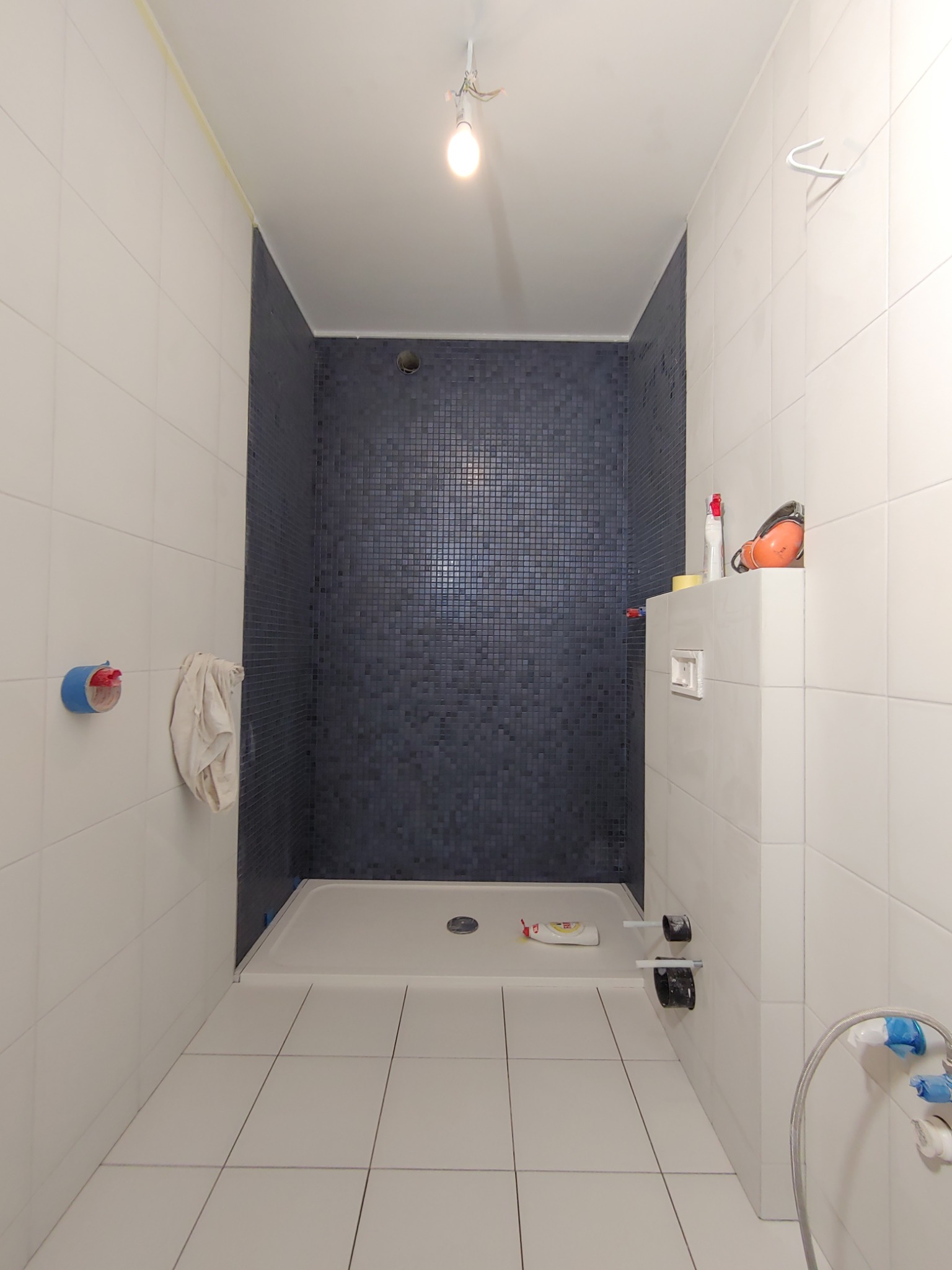 Wnętrze łazienki w trakcie prac wykończeniowych: białe płytki ścienne i podłogowe, kabina prysznicowa z ciemnej mozaiki, widoczne narzędzia i materiały budowlane.