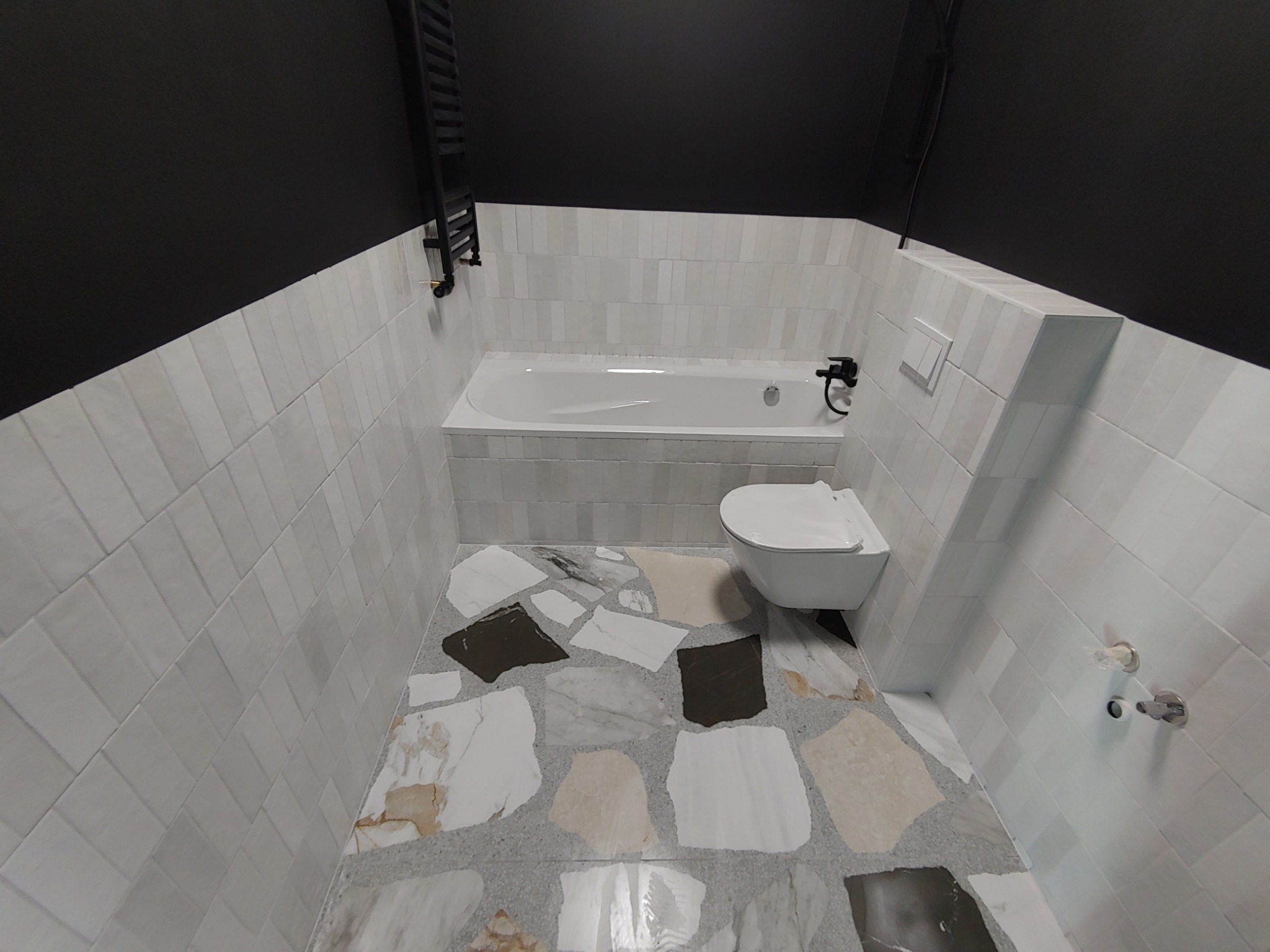 Nowoczesna łazienka z czarnym sufitem, białymi, wąskimi płytkami ściennymi ułożonymi pionowo, mozaikową podłogą z dużych, nieregularnych płyt w odcieniach bieli, szarości i beżu, oraz białą wanną...