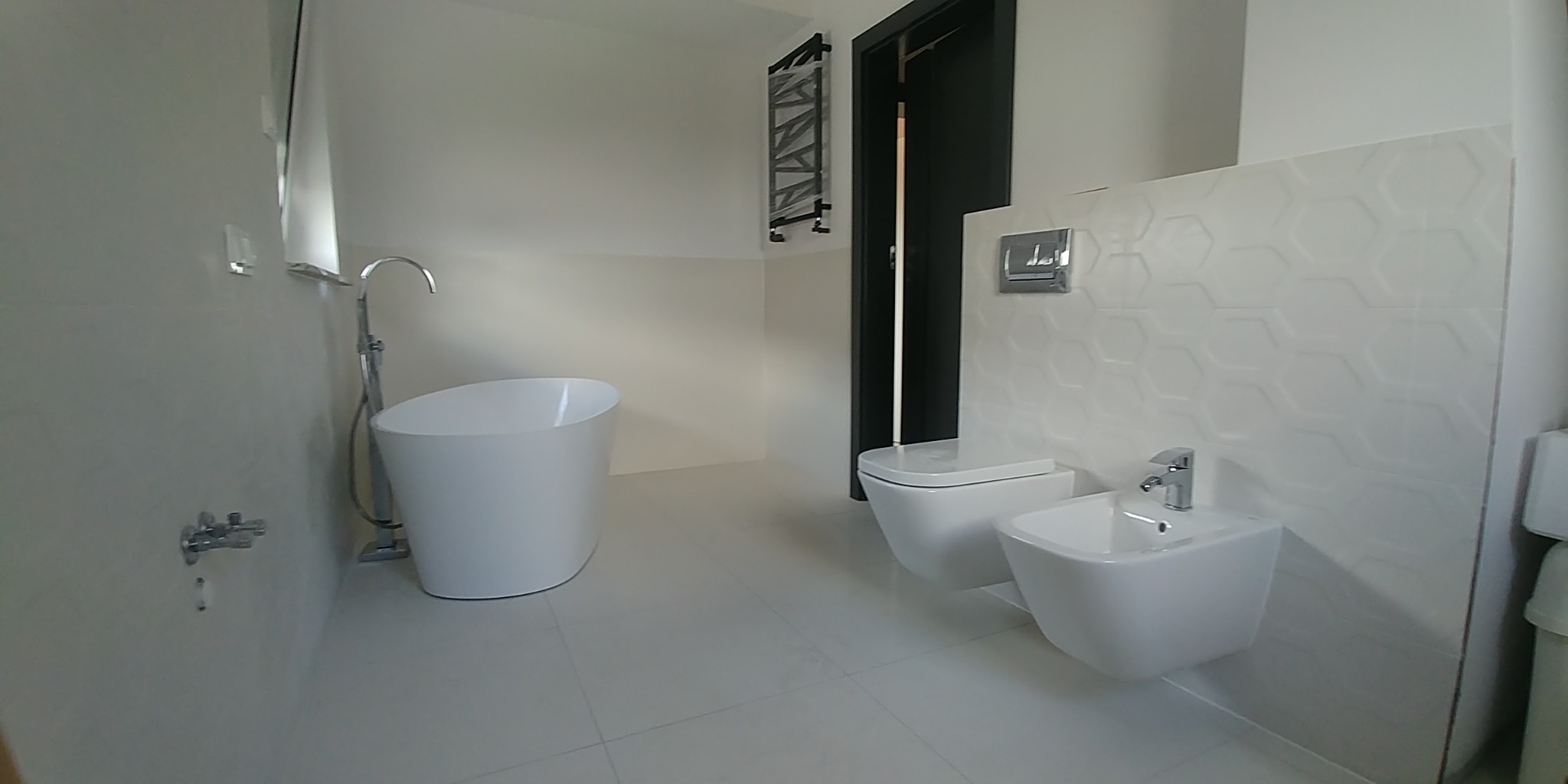 Nowoczesna, minimalistyczna łazienka z wolnostojącą wanną, wiszącą toaletą i bidetem, białe płytki z heksagonalnym wzorem na ścianie, czarne drzwi w tle.