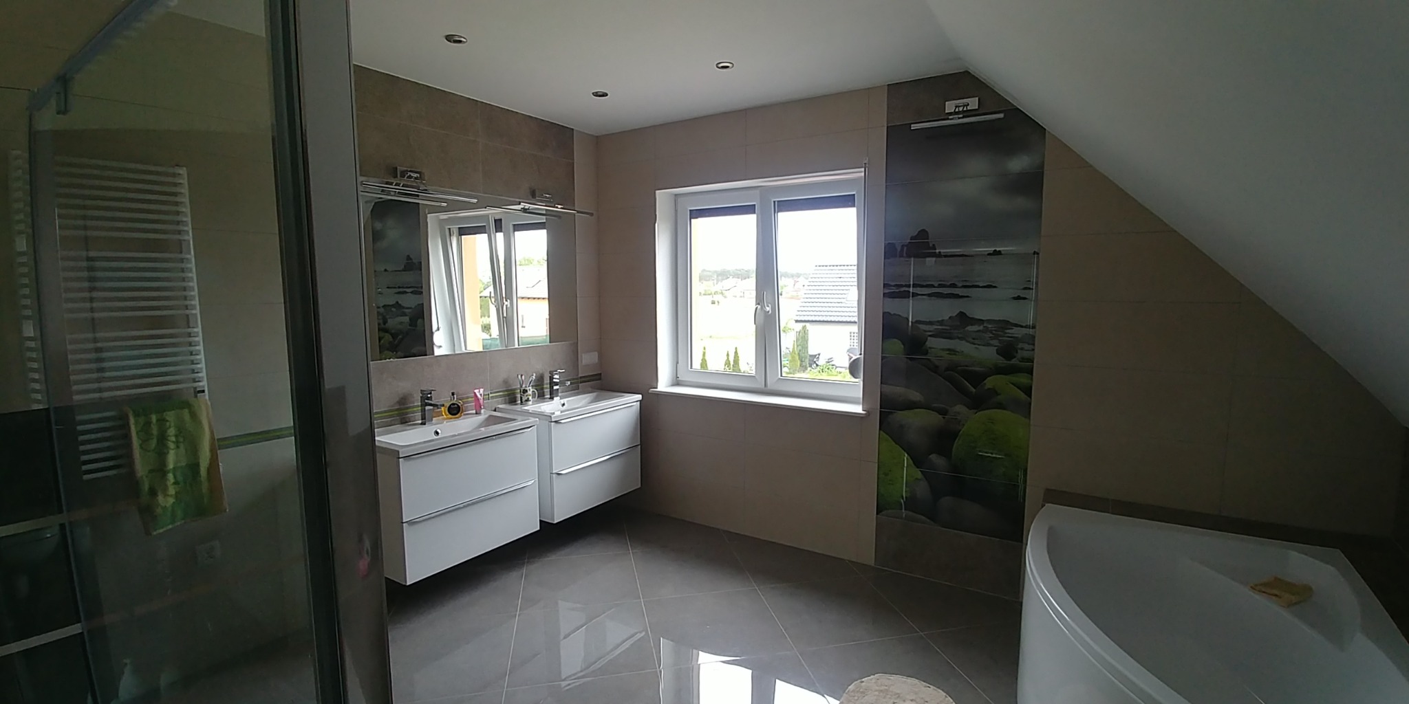 Nowoczesna łazienka na poddaszu z dwoma białymi umywalkami, dużym oknem i dekoracyjną ścianą z motywem morskim.