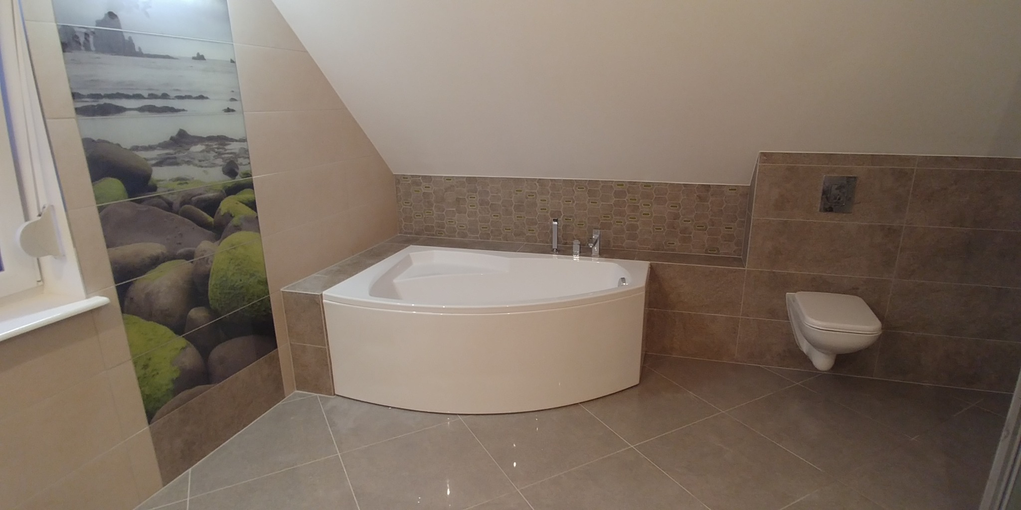 Nowoczesna łazienka na poddaszu z narożną wanną, toaletą podwieszaną i płytkami imitującymi krajobraz morski na ścianie.