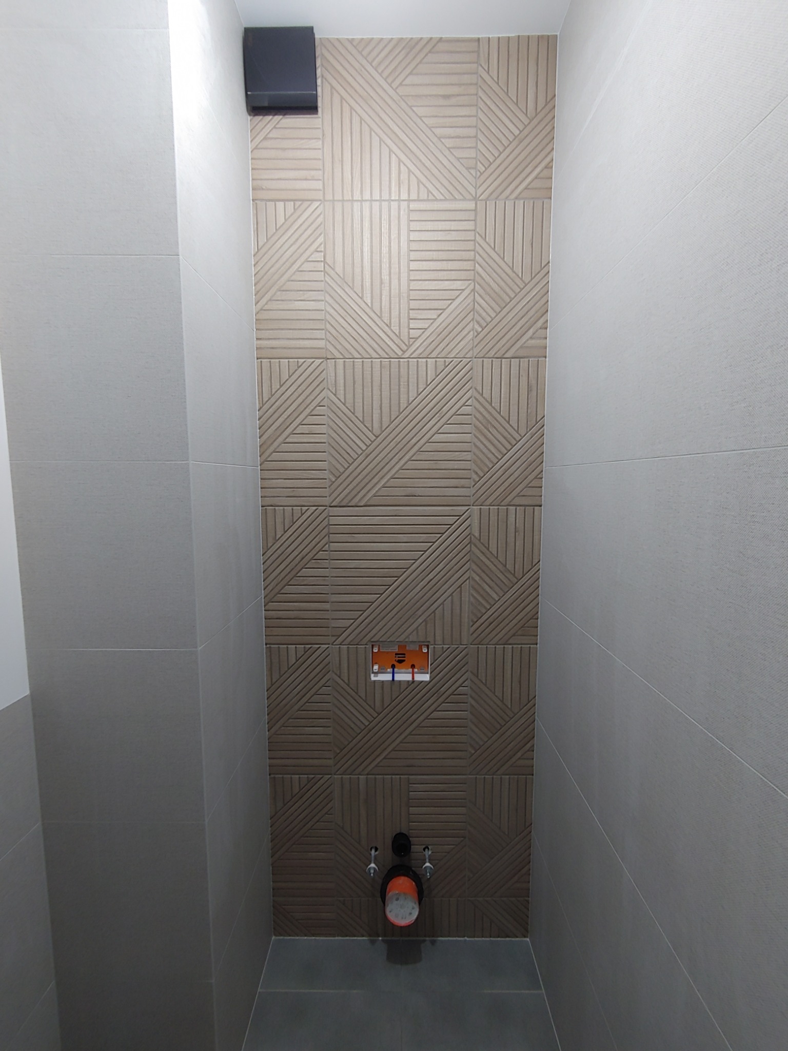 Wąskie ujęcie fragmentu łazienki w trakcie prac wykończeniowych, widoczna ściana z dekoracyjną glazurą w geometryczne wzory, częściowo ułożona, z widocznymi instalacjami hydraulicznymi i wentylacją.