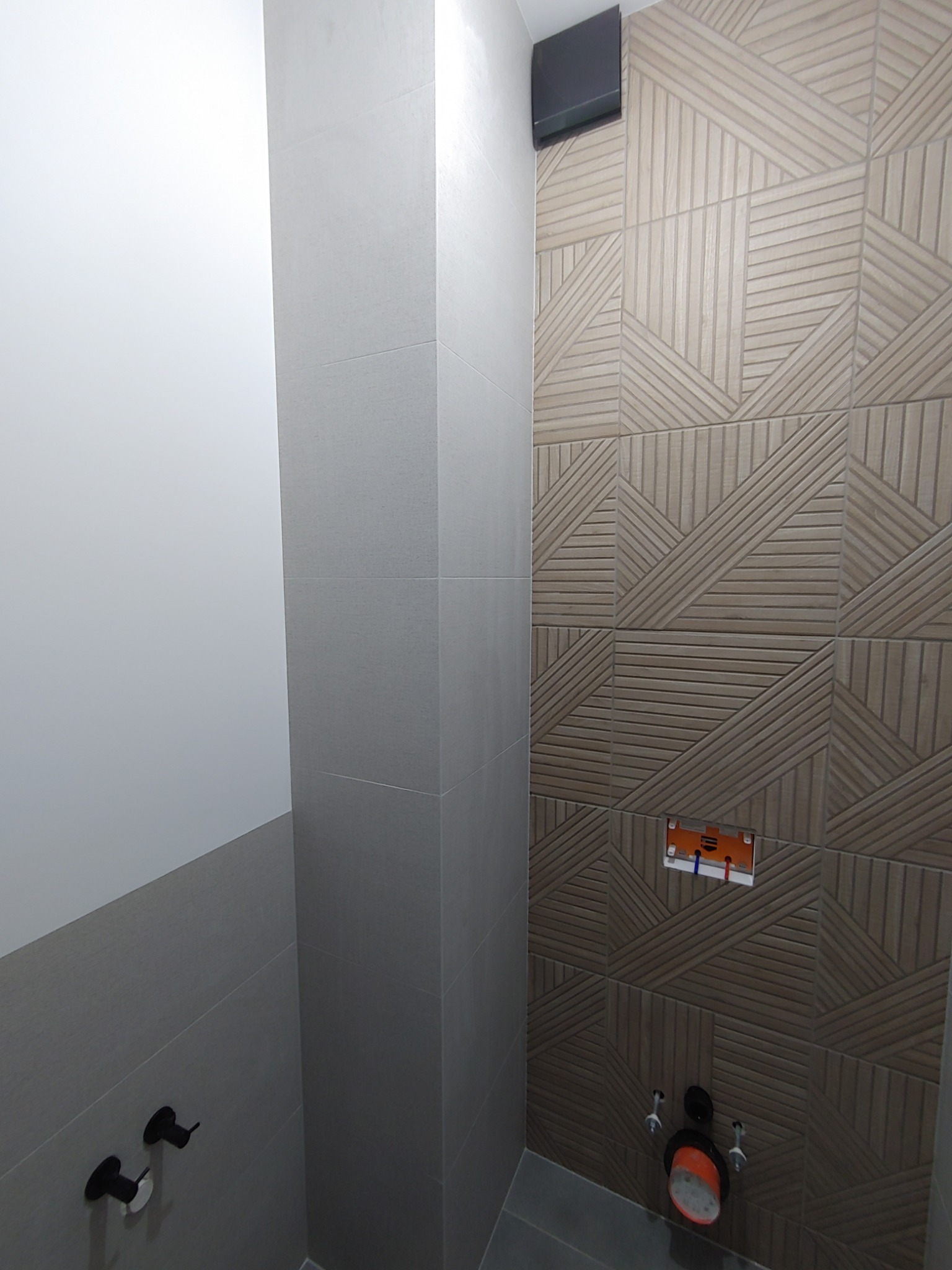 Fragment łazienki w trakcie wykańczania: geometryczne płytki ścienne w odcieniach brązu, szare płytki na ścianie obok, widoczne czarne zawory i elementy instalacji podtynkowych.