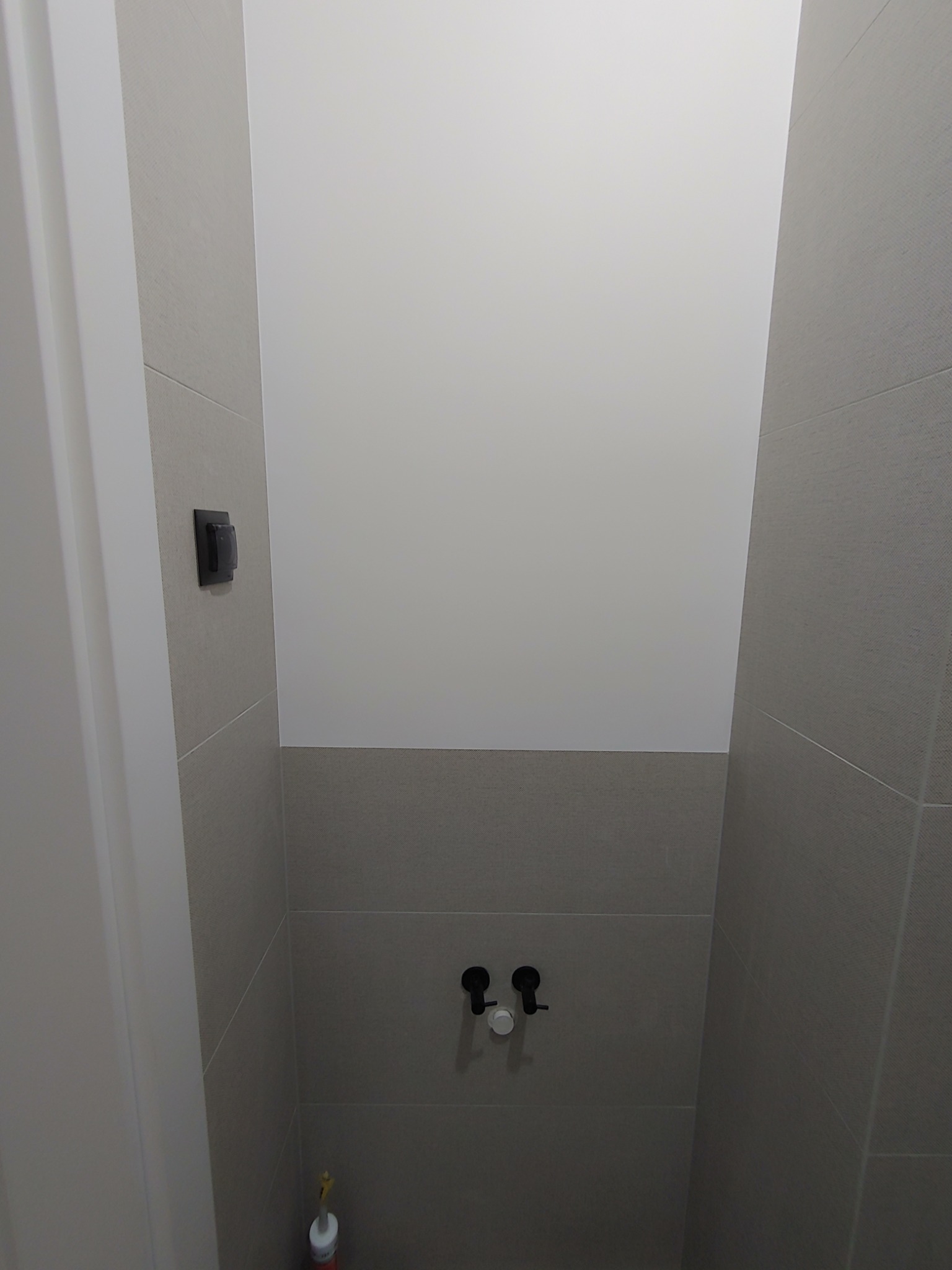 Wnęka w łazience wyłożona szarymi płytkami ceramicznymi, z widocznymi zaworami wodnymi i włącznikiem światła, przygotowana do montażu toalety wiszącej.