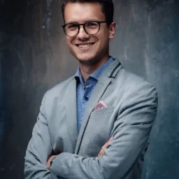 Portret uśmiechniętego mężczyzny w okularach i szarym garniturze z poszetką, stojącego na tle teksturowanej ściany.