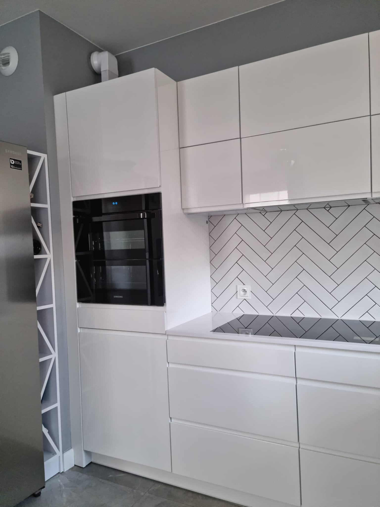 Nowoczesna kuchnia z białymi, lakierowanymi frontami szafek, czarnym piekarnikiem Samsung i płytą indukcyjną, oraz białymi płytkami ułożonymi w jodełkę na ścianie.