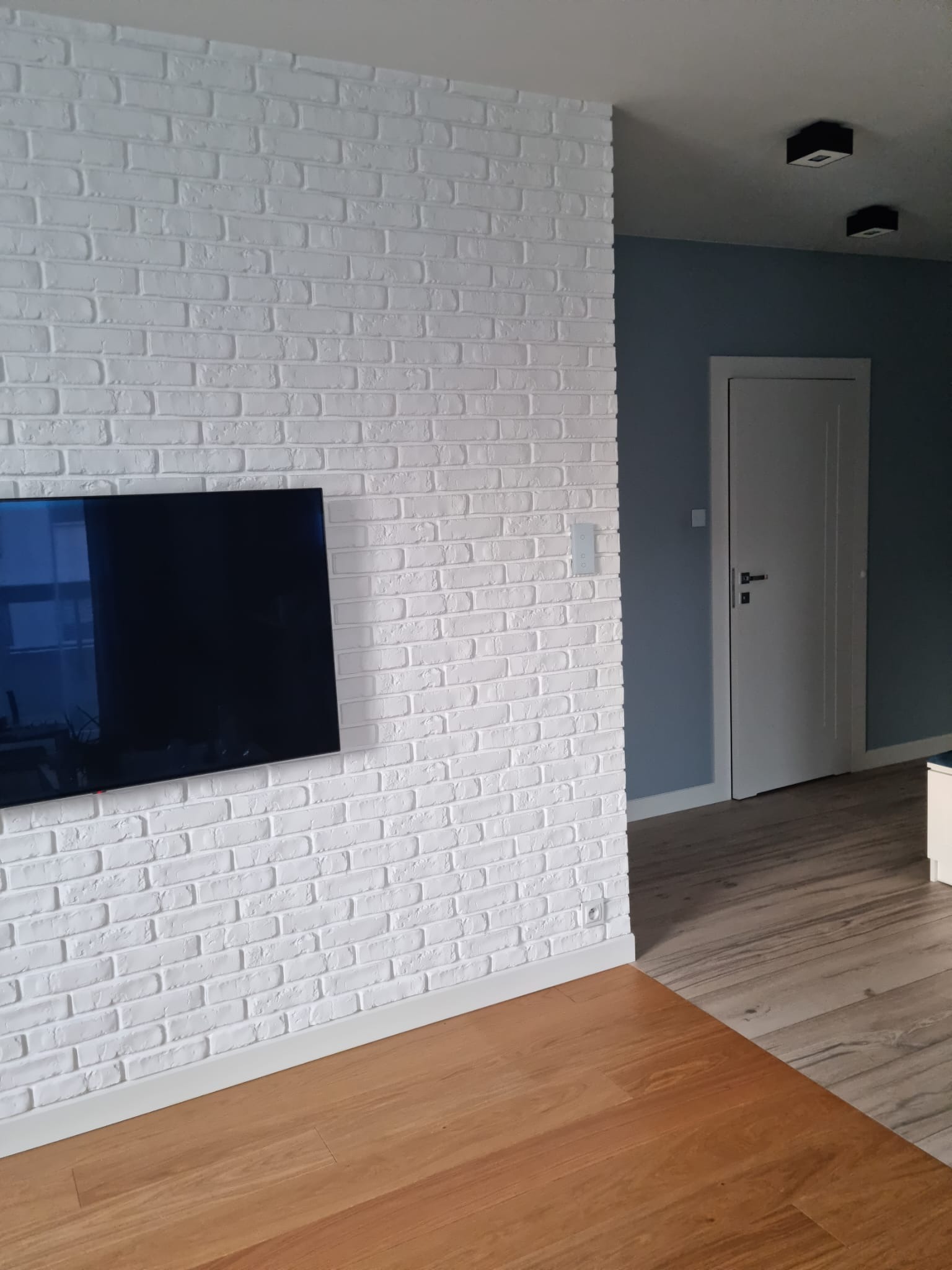 Biała ściana z cegły dekoracyjnej w salonie z telewizorem, panele podłogowe w dwóch odcieniach, widoczny fragment drzwi i oświetlenia sufitowego.