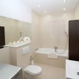 Jasna łazienka z wanną, umywalką i toaletą w zabudowie. Na półce nad toaletą dekoracyjne świece i rolka papieru toaletowego. W lustrze odbicie pralki.