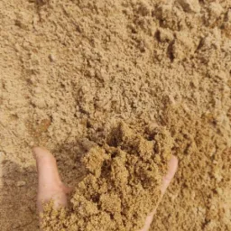 Dłoń trzyma garść drobnego, brązowo-żółtego kruszywa na tle jego większej pryzmy.