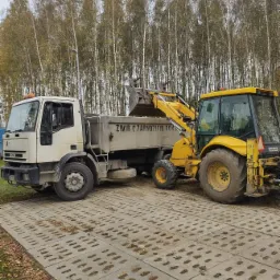 Żółta ładowarka nabiera żwir do skrzyni ładunkowej białej ciężarówki na betonowym podłożu, tło stanowią wysokie drzewa.