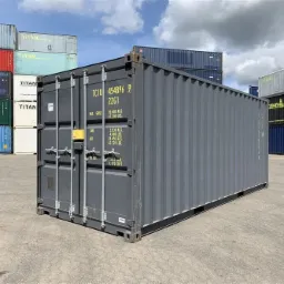 Szary kontener transportowy z widocznymi napisami i numerami, ustawiony na betonowej powierzchni z innymi kontenerami w tle pod pochmurnym niebem.