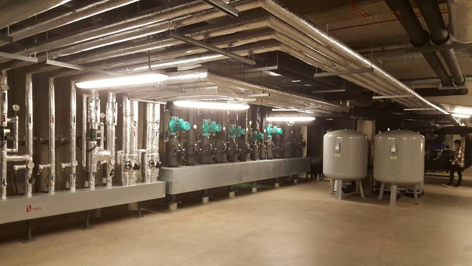Instalacja z rzędami zaworów z zielonymi pokrętłami i izolowanymi rurami pod sufitem, z dwoma szarymi zbiornikami ciśnieniowymi po prawej stronie.