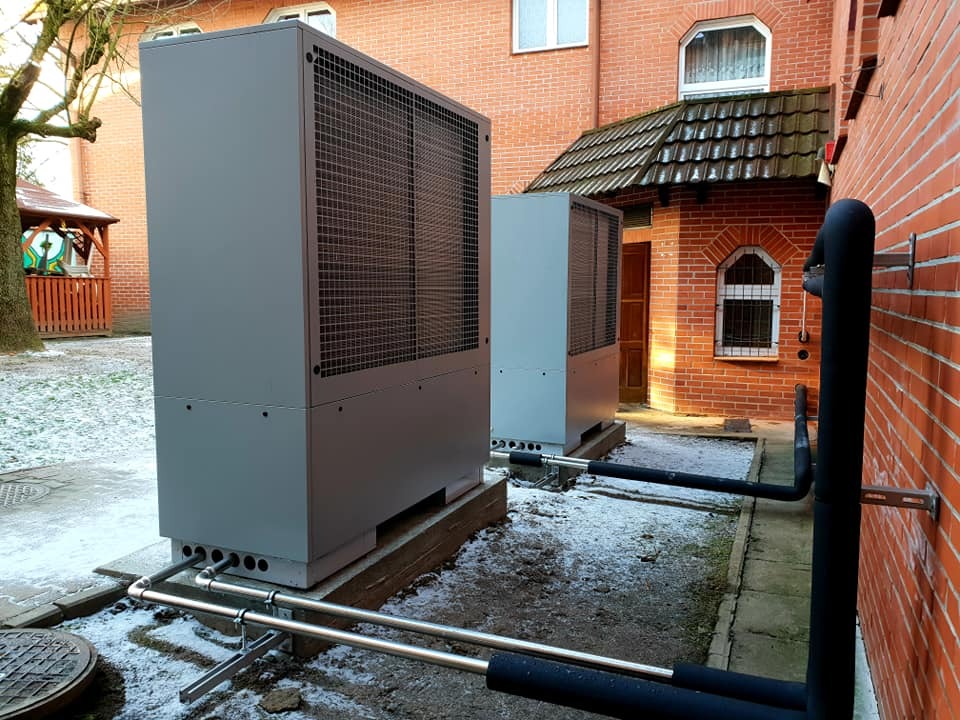 Dwa moduły szarej pompy ciepła zainstalowane na zewnątrz budynku z czerwonej cegły, widoczne rury izolowane termicznie i lekki śnieg na ziemi.