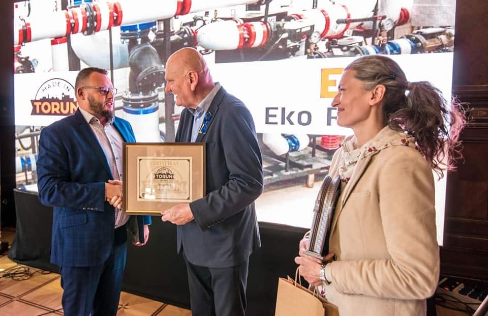 Wręczenie certyfikatu firmie Eko F na tle instalacji z rur, mężczyźni w garniturach ściskają sobie dłonie, kobieta w beżowej marynarce trzyma nagrodę.