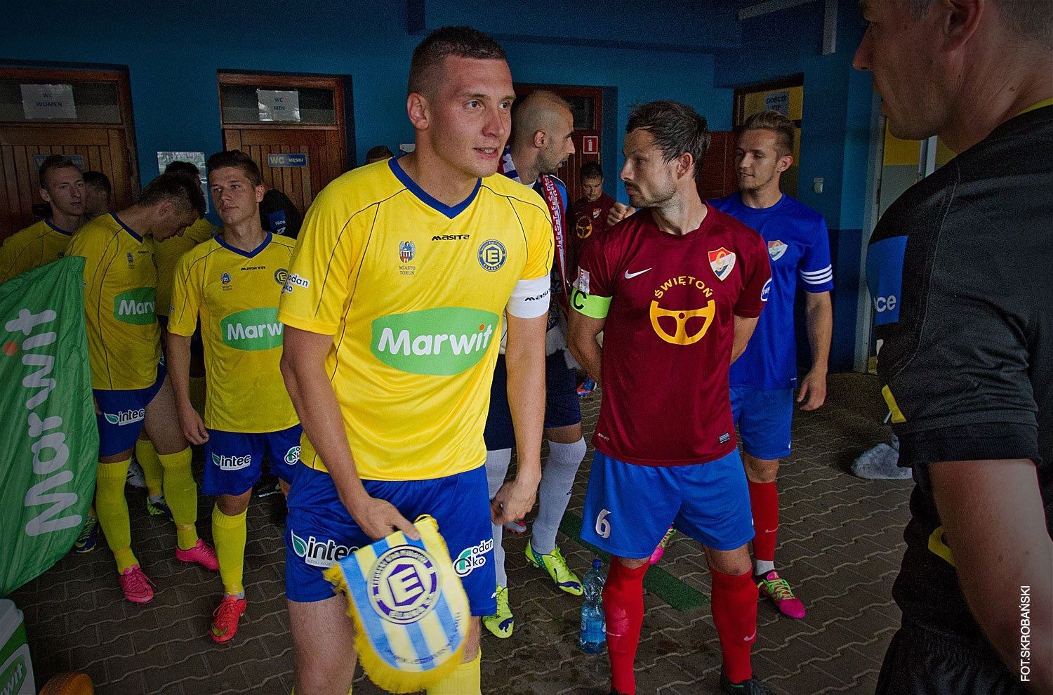 Grupa piłkarzy w szatni przed meczem, ubrani w kolorowe stroje sportowe z widocznymi logotypami sponsorów.